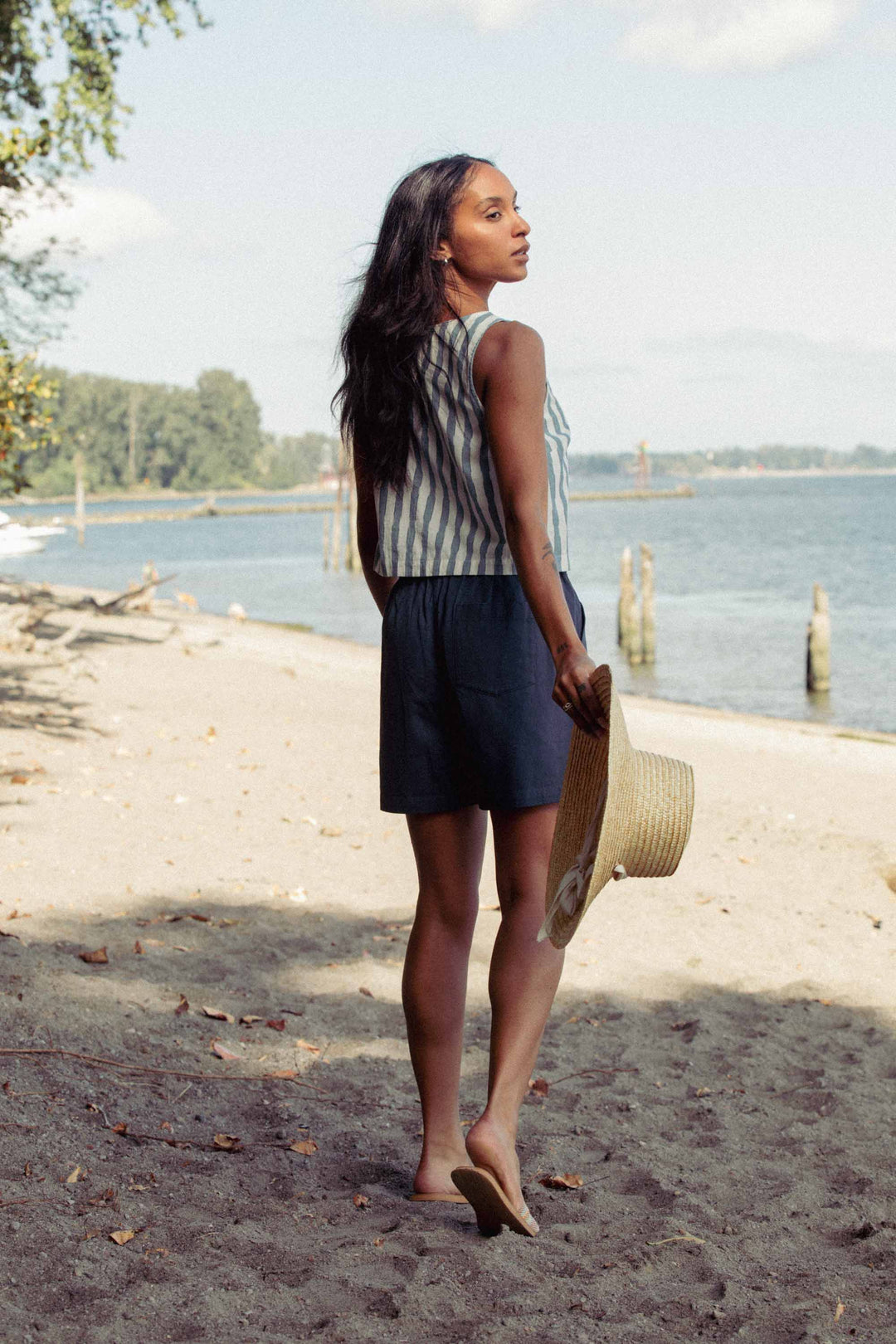 A woman with long hair walks barefoot on a sandy beach, holding a straw hat and wearing the Bridge & Burn Sadie Tank in Beach Stripe with dark shorts. Trees and water can be seen in the background.