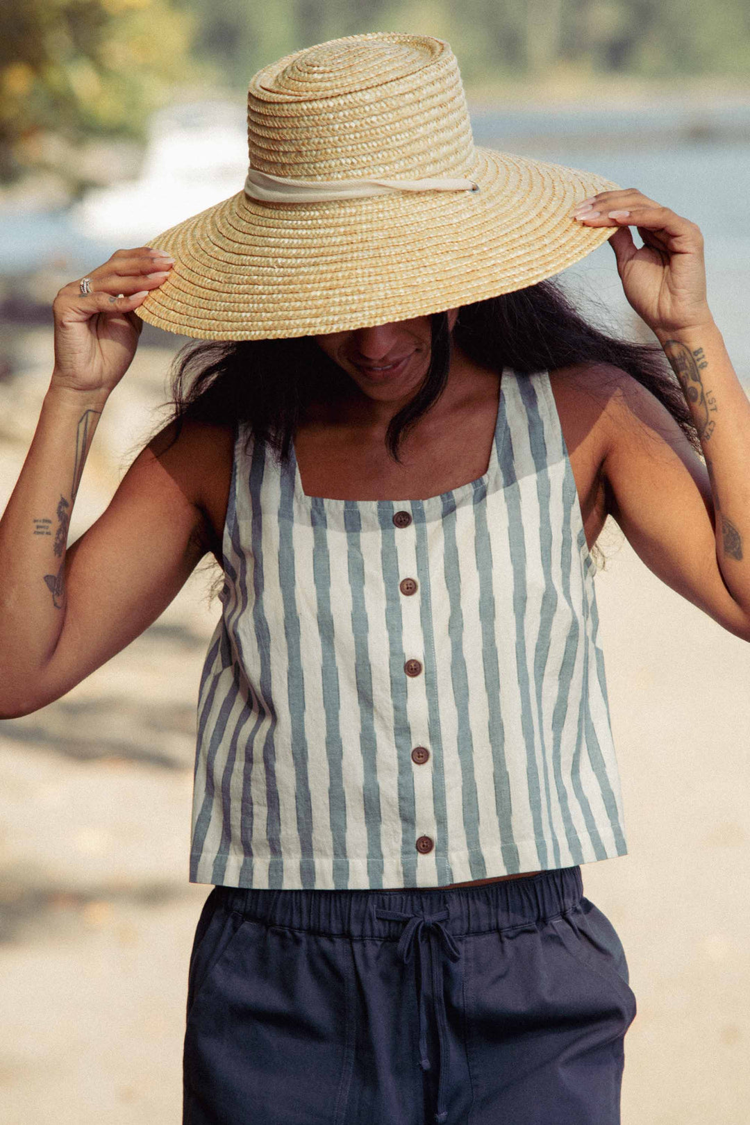 A person stands outdoors in the Bridge & Burn Sadie Tank in Beach Stripe, wearing a wide-brimmed straw hat and dark shorts. They hold the hat’s brim, shading their face, with blurred trees and water providing a tranquil backdrop.
