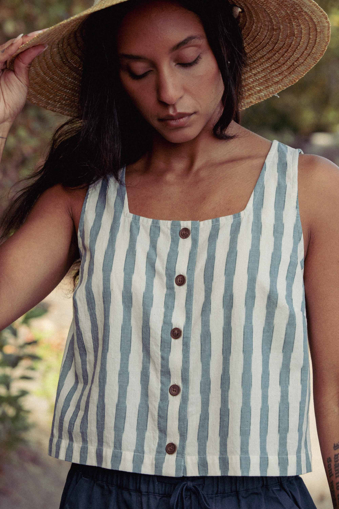 A woman wearing the Bridge & Burn Sadie Tank in Beach Stripe, paired with a wide-brimmed straw hat, stands outdoors and looks down with a calm expression.