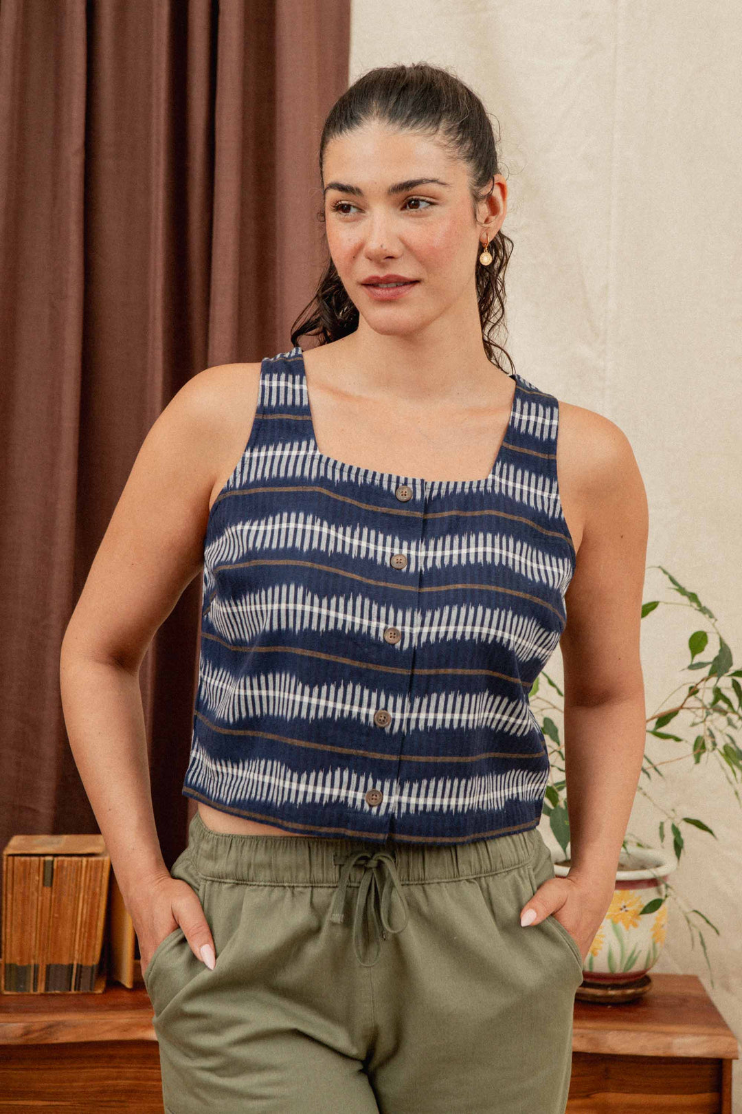 A woman with dark hair tied back wears the Bridge & Burn Sadie Tank in Dusk Mirage and olive pants, standing indoors with her hands in her pockets near plants and wooden decor.