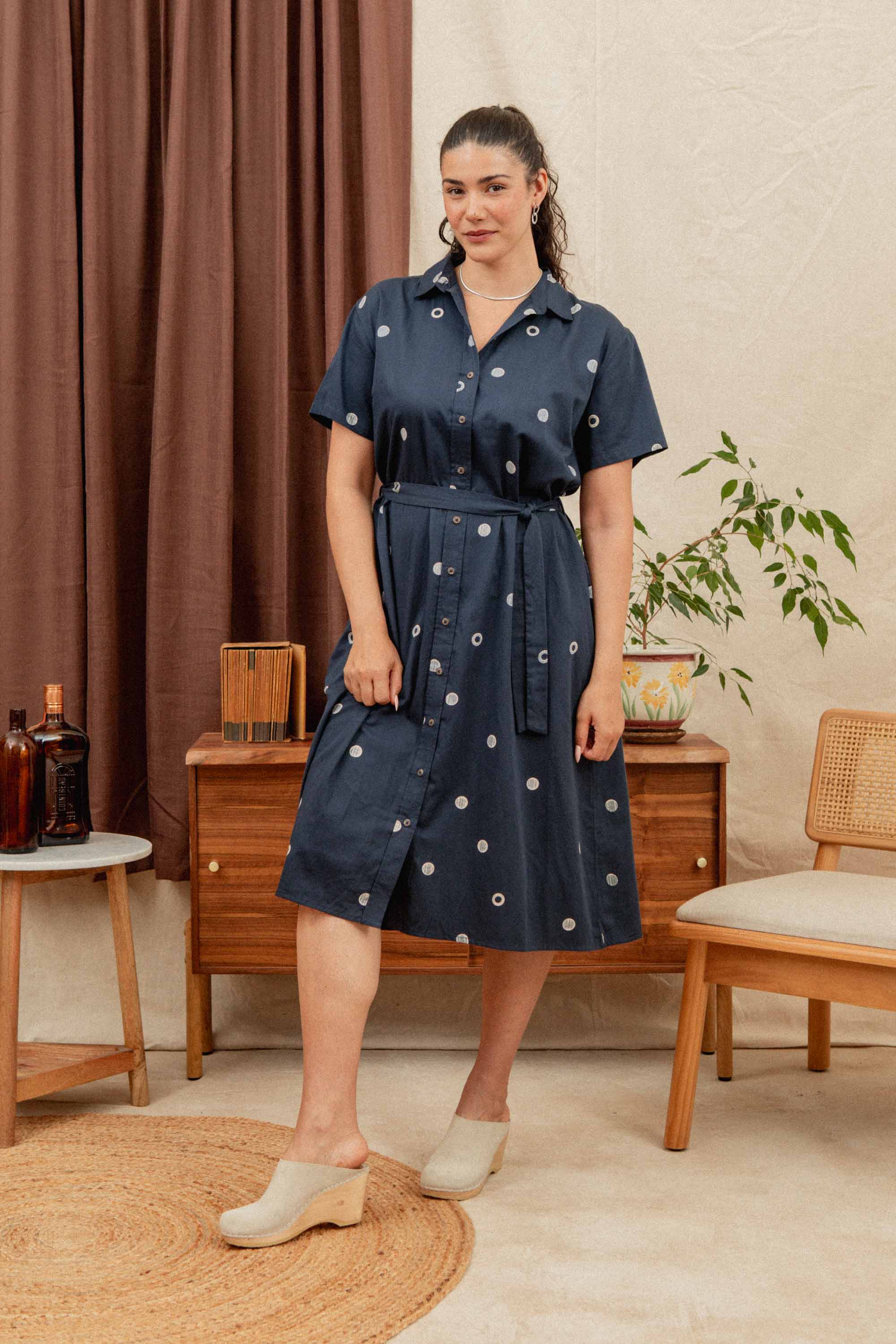 A woman wears the Bridge & Burn Sylvie Dress in Lunar Polka—navy blue organic cotton with white circles—beside mid-century furniture, a plant, and brown curtains. Her hair is tied back and she pairs it with cream-colored shoes.