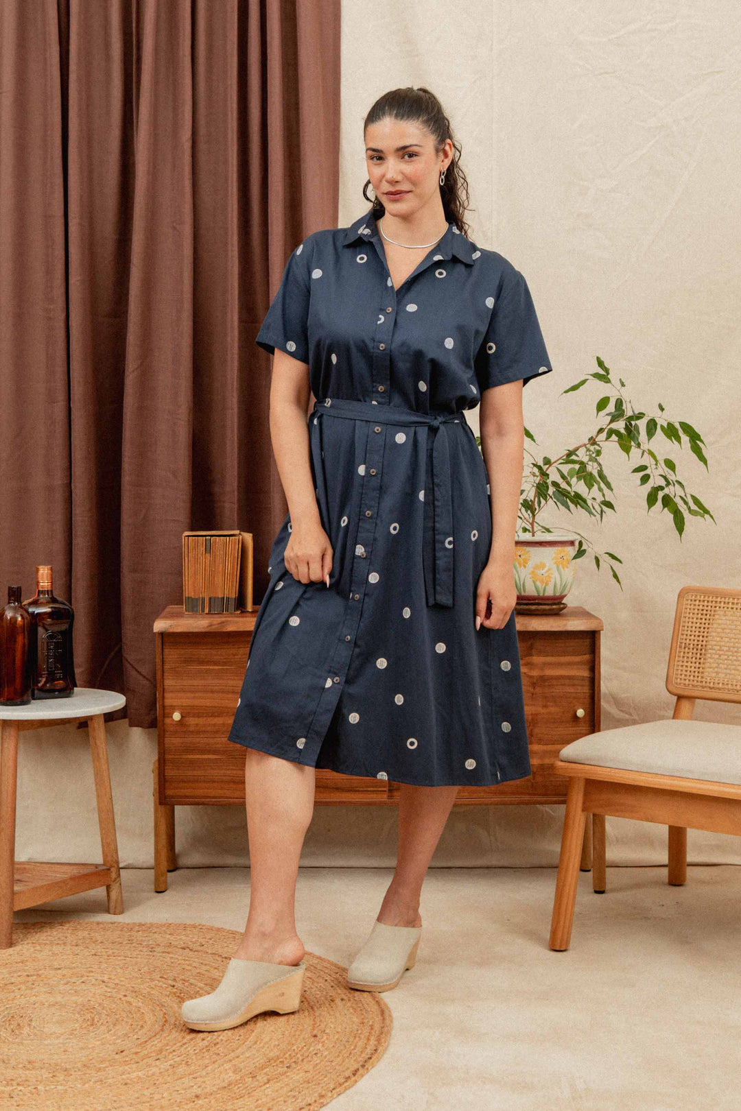 A woman wears the Bridge & Burn Sylvie Dress in Lunar Polka—navy blue organic cotton with white circles—beside mid-century furniture, a plant, and brown curtains. Her hair is tied back and she pairs it with cream-colored shoes.