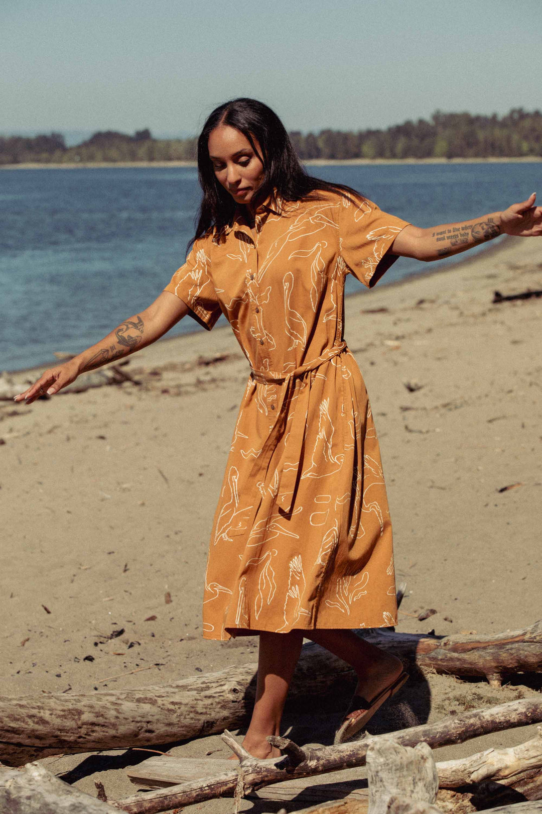 A woman wears the Bridge & Burn Sylvie Dress in Shoreflight while walking barefoot along a sandy beach with driftwood, calm water, and trees in the background.