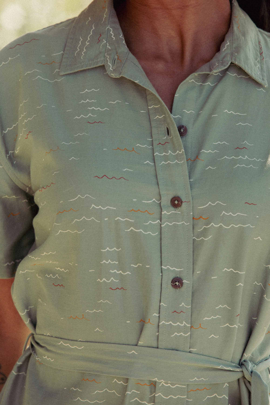 A close-up of a person wearing the Bridge & Burn Sylvie Dress in Teal Tide Waves, featuring short sleeves, a fabric waist belt, and a colorful wavy line pattern. The image highlights the relaxed upper torso fit.