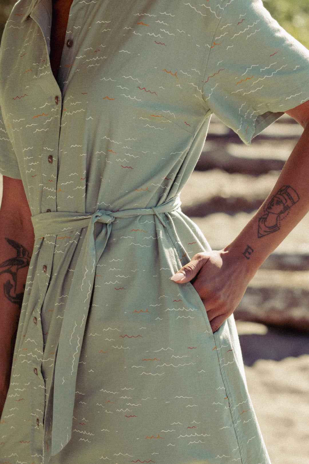 Outdoors, a person wears the Bridge & Burn Sylvie Dress in Teal Tide Waves—a light green organic cotton shirtdress with short sleeves, waist tie, pockets, and subtle wavy patterns. Forearm tattoos show as their hand rests in a pocket; logs blur behind.