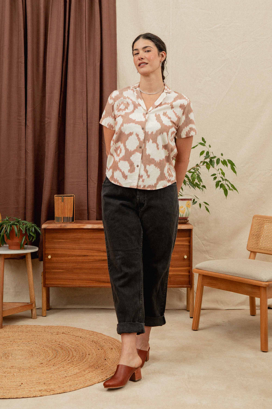 A woman stands indoors on a woven rug, wearing the Bridge & Burn Wren Shirt in Pop Leaf—a retro-inspired, lightweight cotton top—paired with dark cuffed pants and brown heeled shoes. Wooden furniture and houseplants decorate the background.
