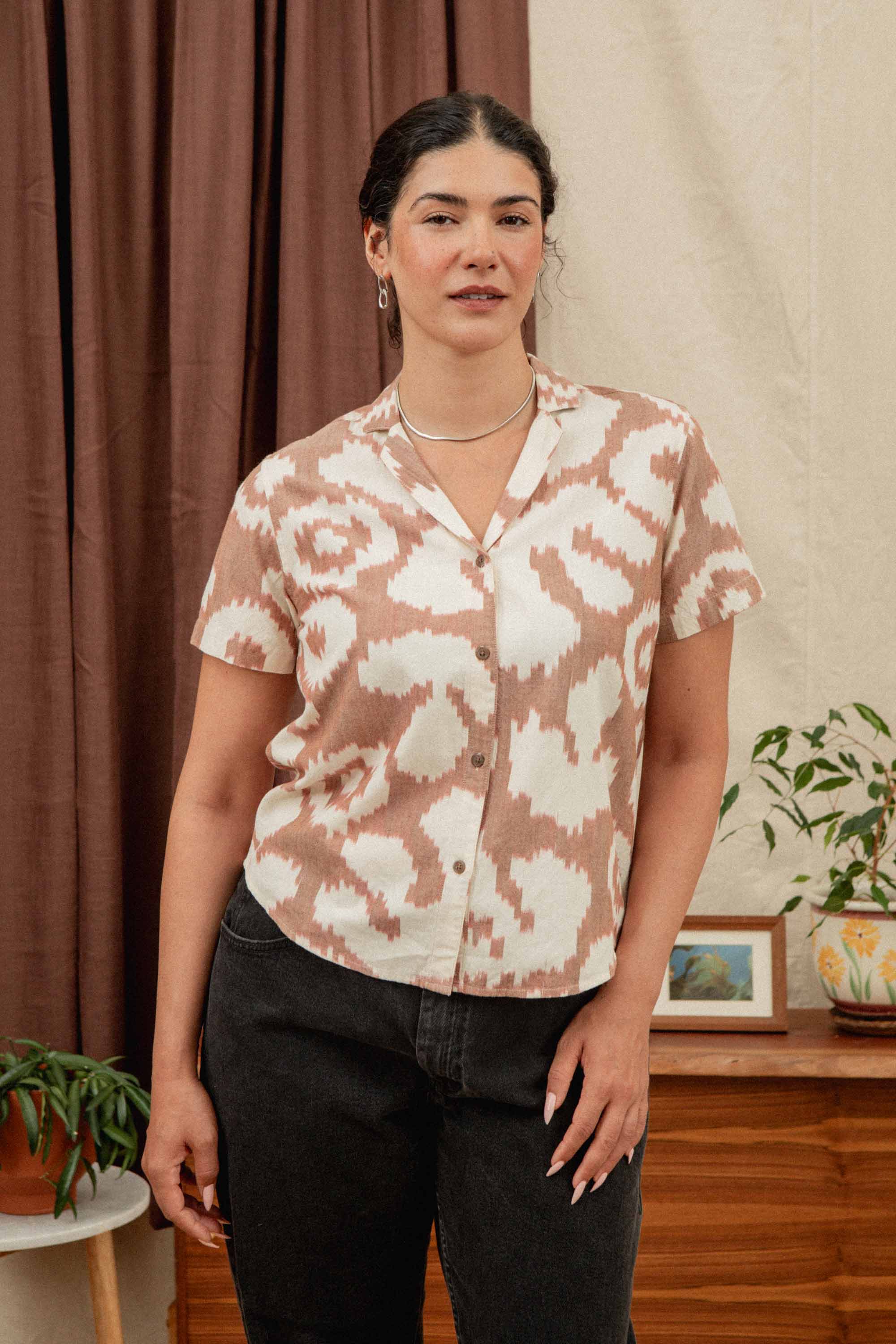 A woman with dark hair wears the Bridge & Burn Wren Shirt in Pop Leaf, a retro-inspired beige and white cotton blouse, paired with dark pants. She stands indoors near houseplants, a wooden cabinet, and cream-brown curtains—ideal for summer comfort.