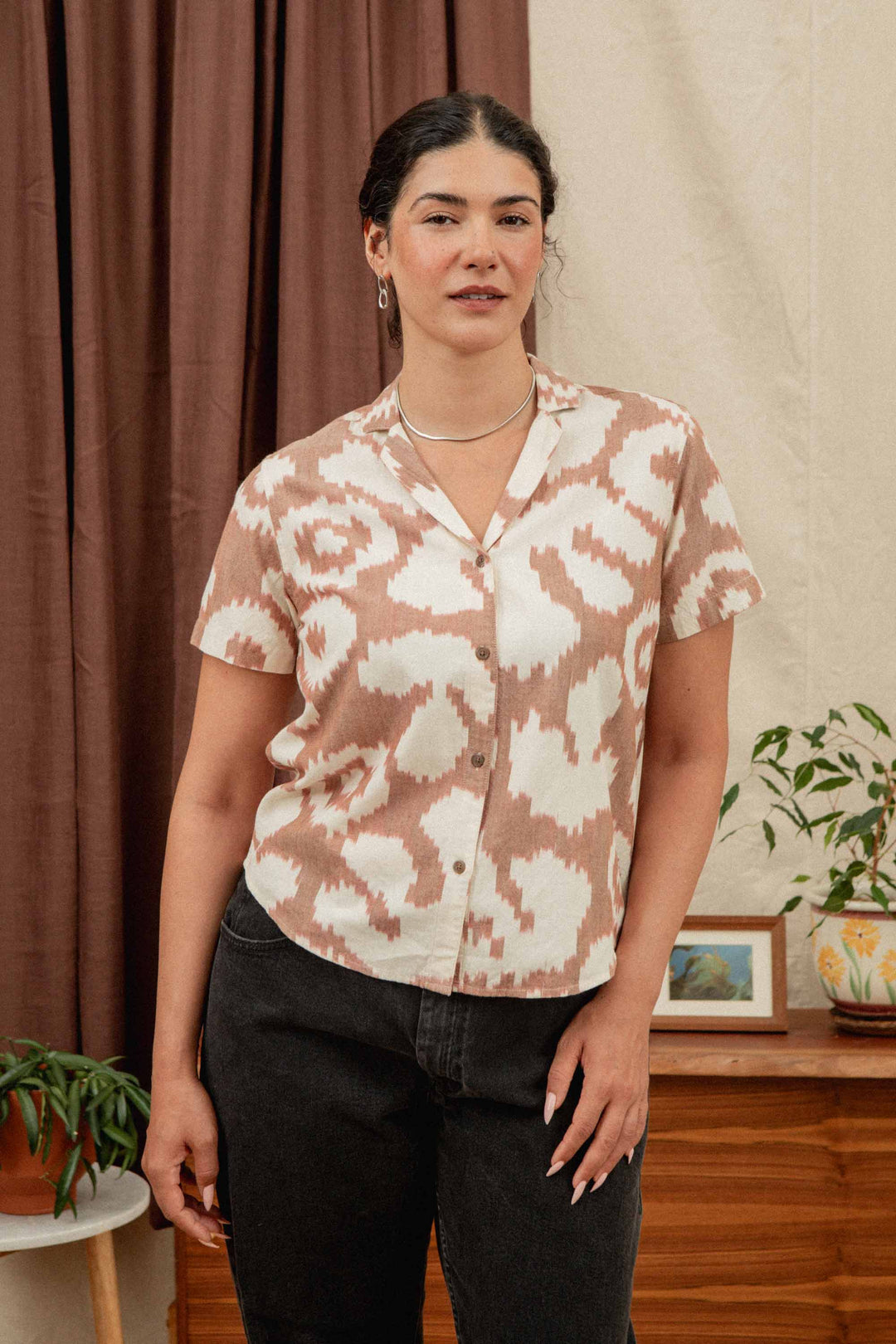 A woman with dark hair wears the Bridge & Burn Wren Shirt in Pop Leaf, a retro-inspired beige and white cotton blouse, paired with dark pants. She stands indoors near houseplants, a wooden cabinet, and cream-brown curtains—ideal for summer comfort.