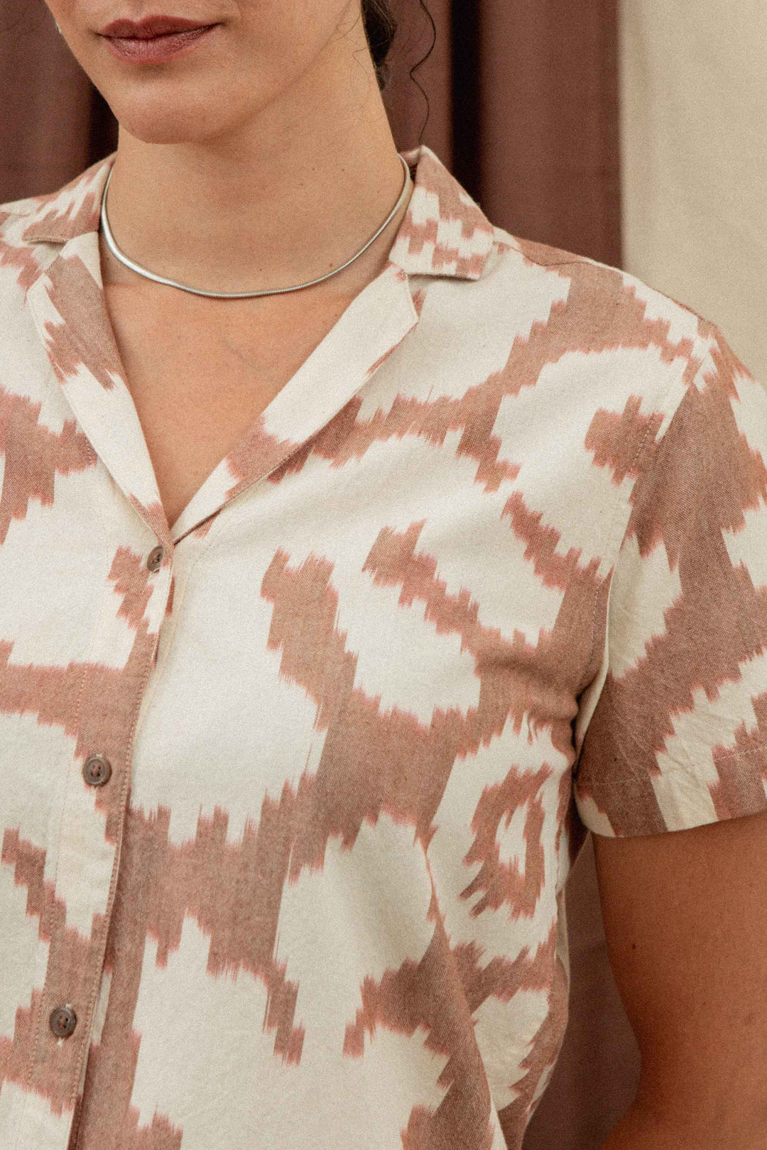 A woman in a Bridge & Burn Wren Shirt / Pop Leaf, featuring a pink and white geometric pattern, stands before a neutral background. The short-sleeve top is perfect for summer comfort, with only her lower face and upper torso visible.