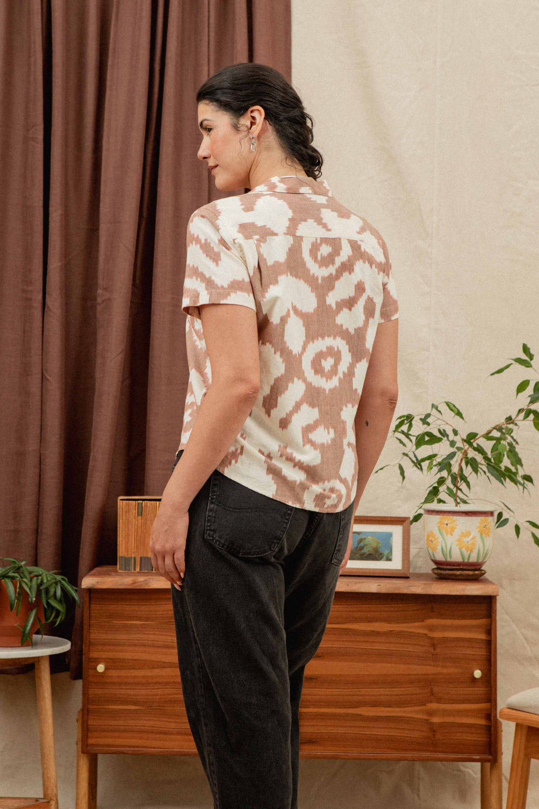 A woman with dark hair in a low bun stands indoors, facing away. She wears the Bridge & Burn Wren Shirt in Pop Leaf cotton and black jeans. Houseplants, a wooden sideboard, and brown curtains are in the background.