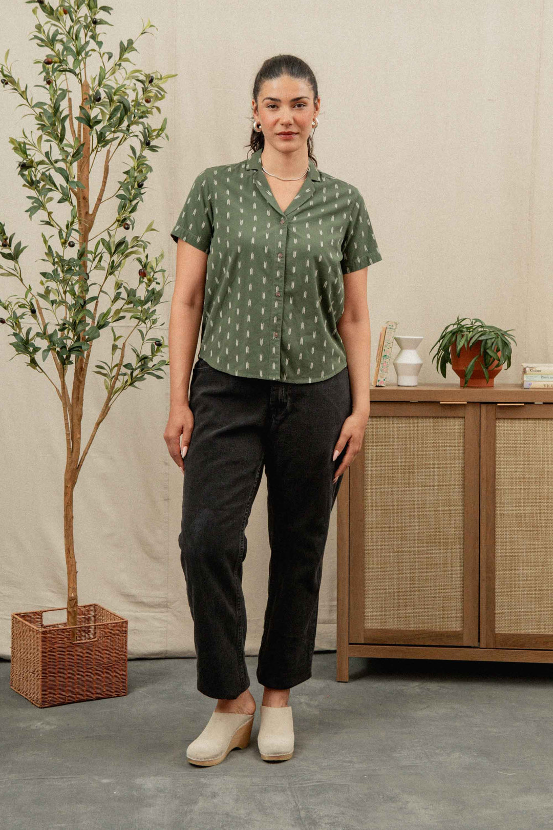 A woman with dark hair tied back wears the Bridge & Burn Wren Shirt in Thyme Arrows, paired with black pants and light-colored shoes. She stands indoors beside a leafy potted plant and a wooden cabinet decorated with ornaments.