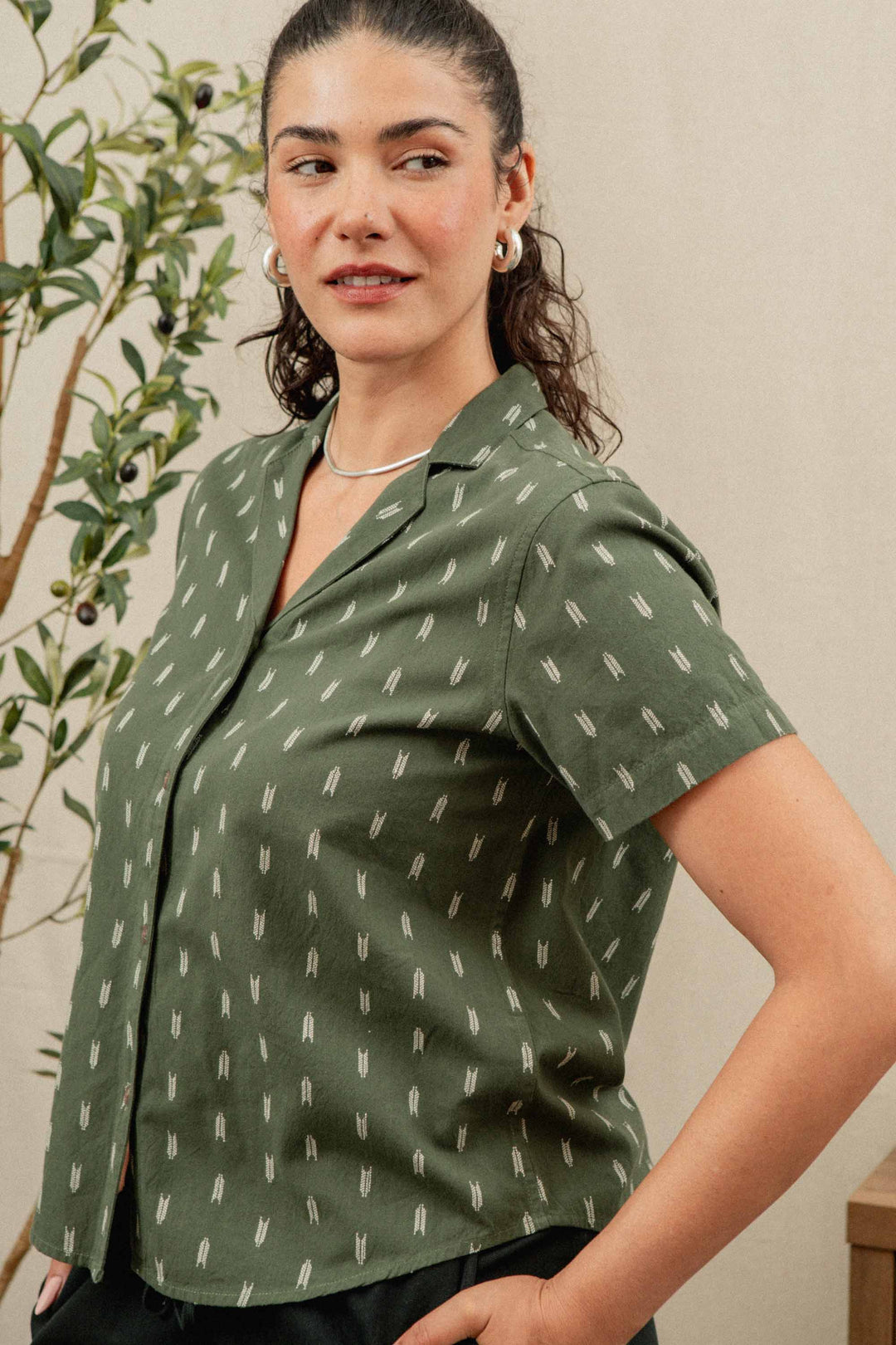 A woman with curly hair, wearing the Bridge & Burn Wren Shirt in the Thyme Arrows pattern and jewelry, stands sideways near a leafy indoor plant, looking slightly over her shoulder.