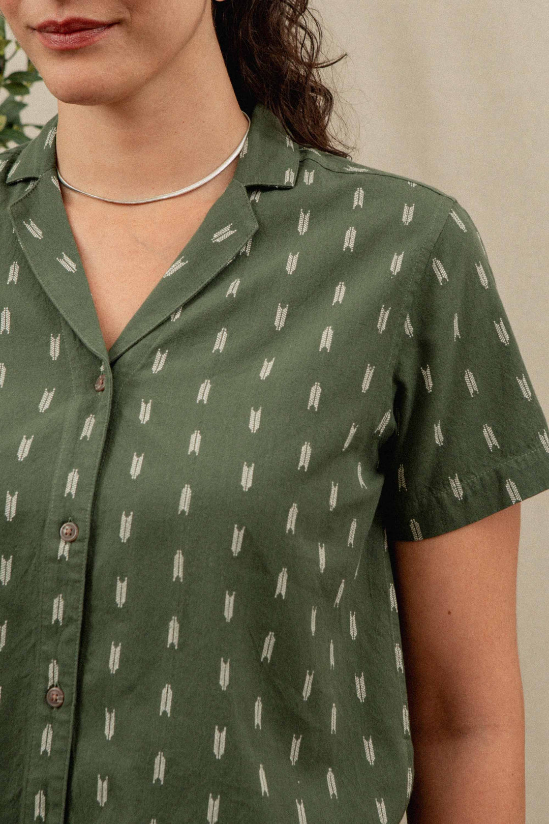A woman wears the Bridge & Burn Wren Shirt in Thyme Arrows—a relaxed-fit, short-sleeved green button-up with a white arrow pattern and lightweight cotton—paired with a silver choker; only her mouth, neck, and torso are visible.