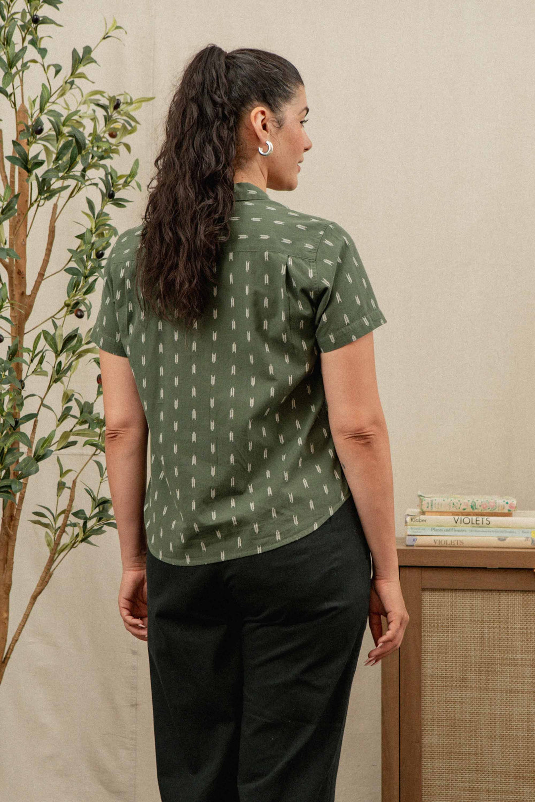 A woman with long dark hair in a ponytail wears the Bridge & Burn Wren Shirt in Thyme Arrows—a short-sleeved, relaxed-fit green cotton top—with black pants, standing near a small table with books and a potted plant.
