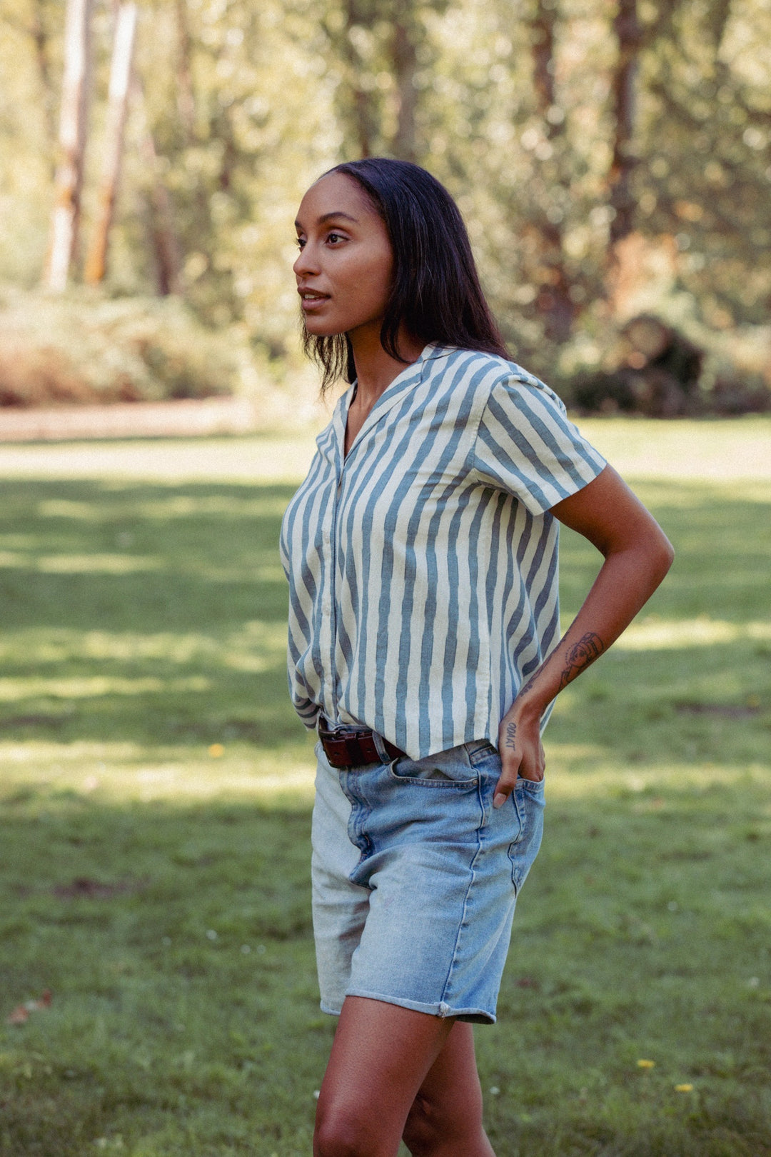 A woman stands on grass under the sun, wearing the Bridge & Burn Wren Shirt in Beach Stripe—white with blue stripes—and denim shorts. She has long dark hair and looks to the side, with trees in the background.