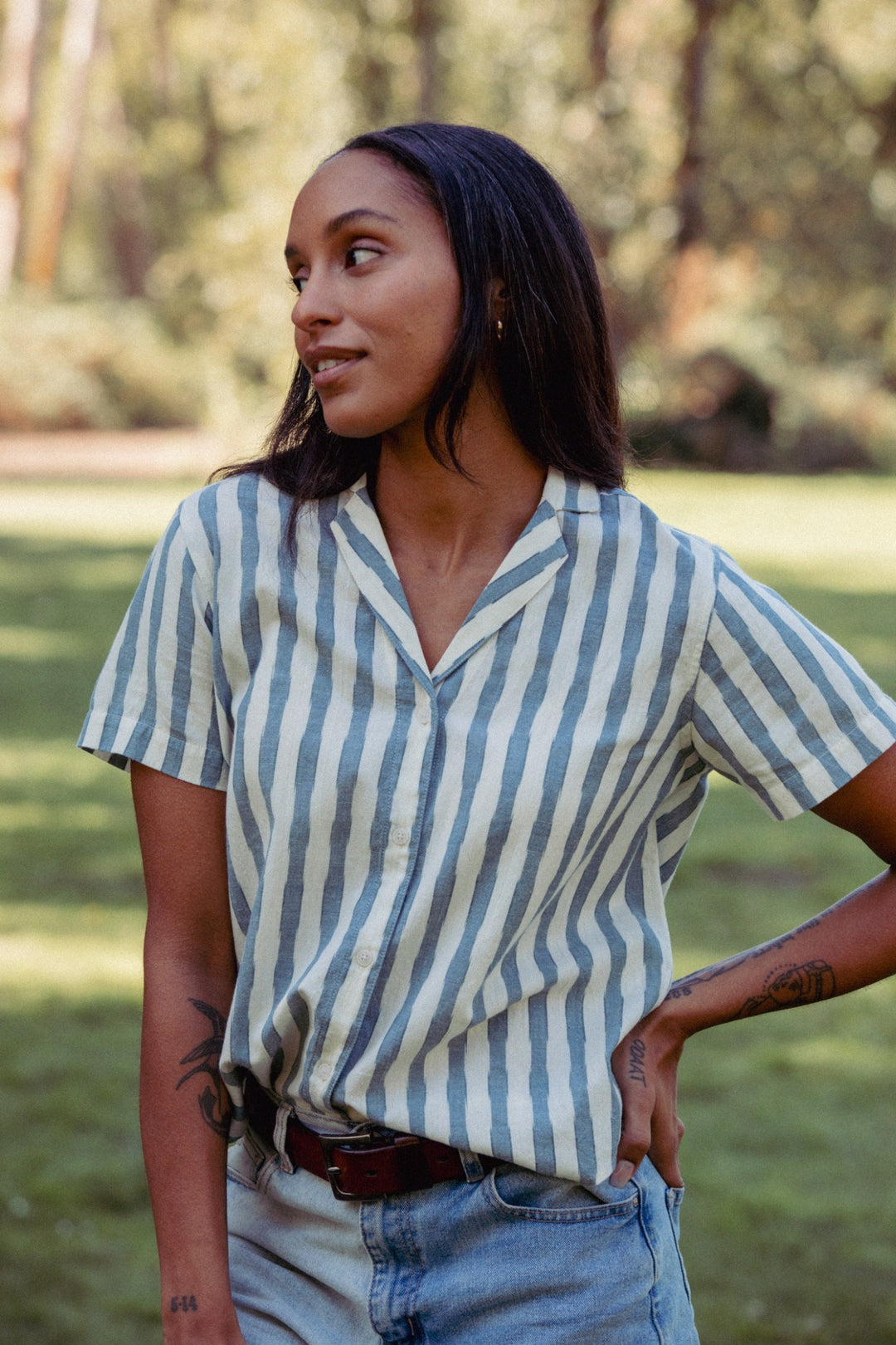 A woman with long dark hair stands on grass, wearing the Bridge & Burn Wren Shirt in Beach Stripe—a relaxed, blue-and-white striped button-up—and light denim jeans. She looks to the side with one hand on her hip. Trees blur in the background.