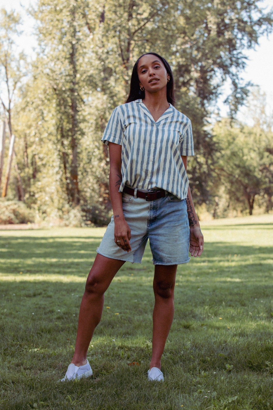 A woman stands on grass in a sunlit park, wearing the Bridge & Burn Wren Shirt in Beach Stripe, styled with denim shorts and white shoes. Trees and greenery fill the background on a clear day.