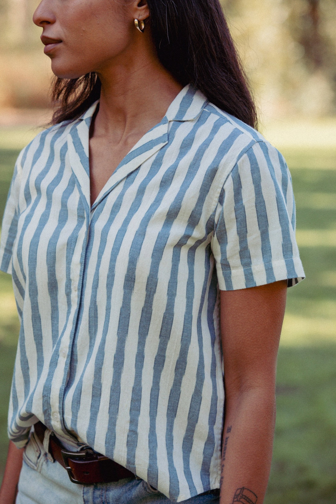 A person outdoors wears the Bridge & Burn Wren Shirt in Beach Stripe, stylishly tucked into light blue jeans. The relaxed, retro-inspired shirt pairs with gold hoop earrings, visible left forearm tattoos, and long dark hair.
