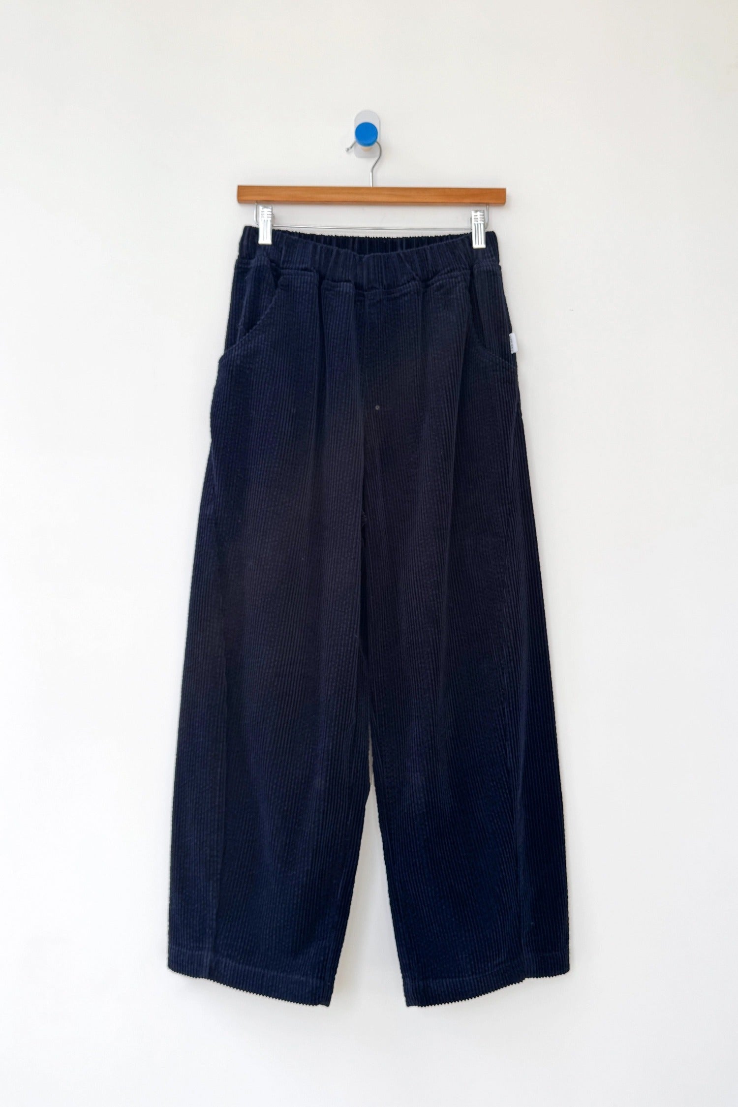 Le Bon Shoppe's Arc Pant in Midnight Corduroy features a wide-leg, high-waist design with an elastic waistband and cropped legs, shown on a wooden hanger against a plain white background.