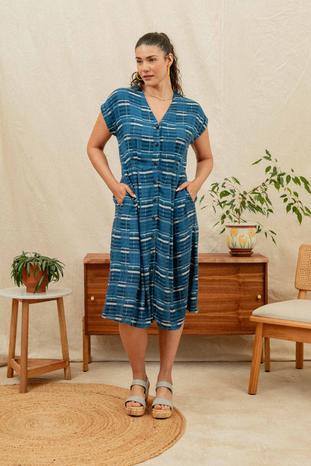 A woman wears the Bridge & Burn Eileen Dress in Stormcrest Stripe, standing indoors with hands in her pockets and sandals on, posed before a wooden sideboard with potted plants. A neutral background enhances the cozy scene.