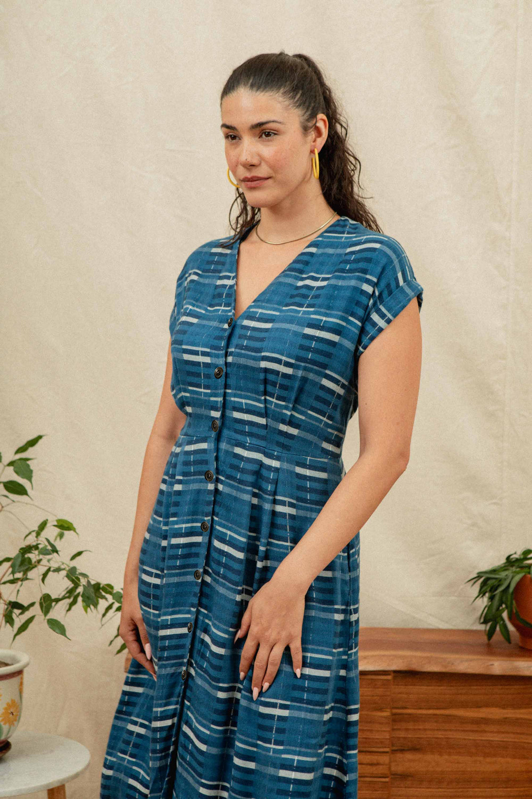 A woman with long brown hair in a ponytail wears the Bridge & Burn Eileen Dress in Stormcrest Stripe, paired with yellow hoop earrings, standing indoors near wooden furniture and potted plants.