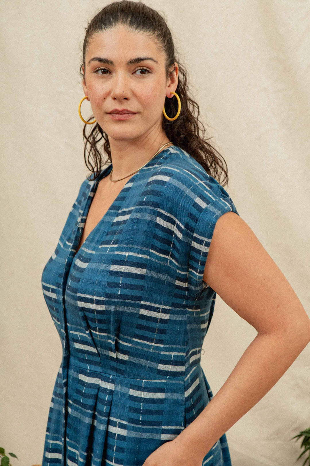 A woman with long, curly hair tied back, wearing large yellow hoop earrings and the Bridge & Burn Eileen Dress in Stormcrest Stripe, stands against a light beige background, gazing slightly to the side with a neutral expression.