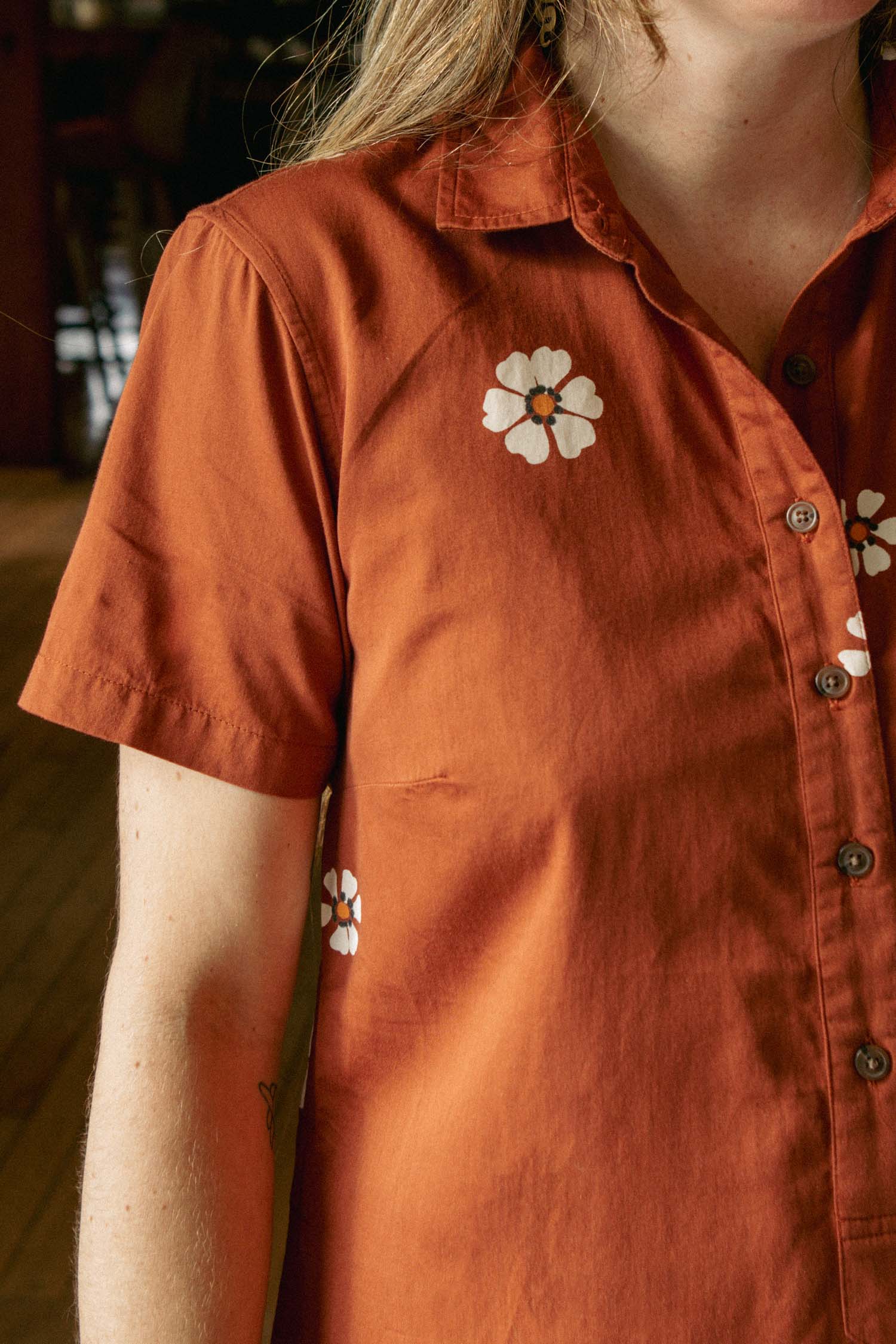 Someone wearing the Bridge & Burn Delia Dress in Redstone Bloom—a short-sleeve shirt dress with a white floral print—stands indoors, their face partially out of frame.