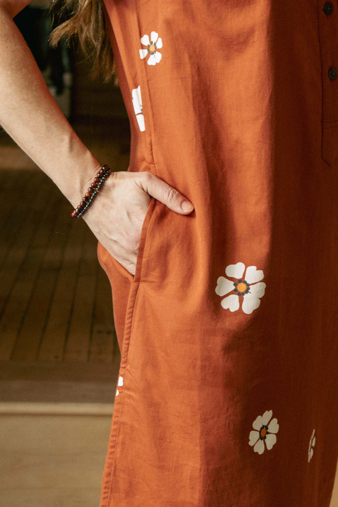 A person wears the Bridge & Burn Delia Dress in Redstone Bloom, featuring orange with white flower prints. One hand is in the pocket, and two beaded bracelets are visible against a wooden floor backdrop.