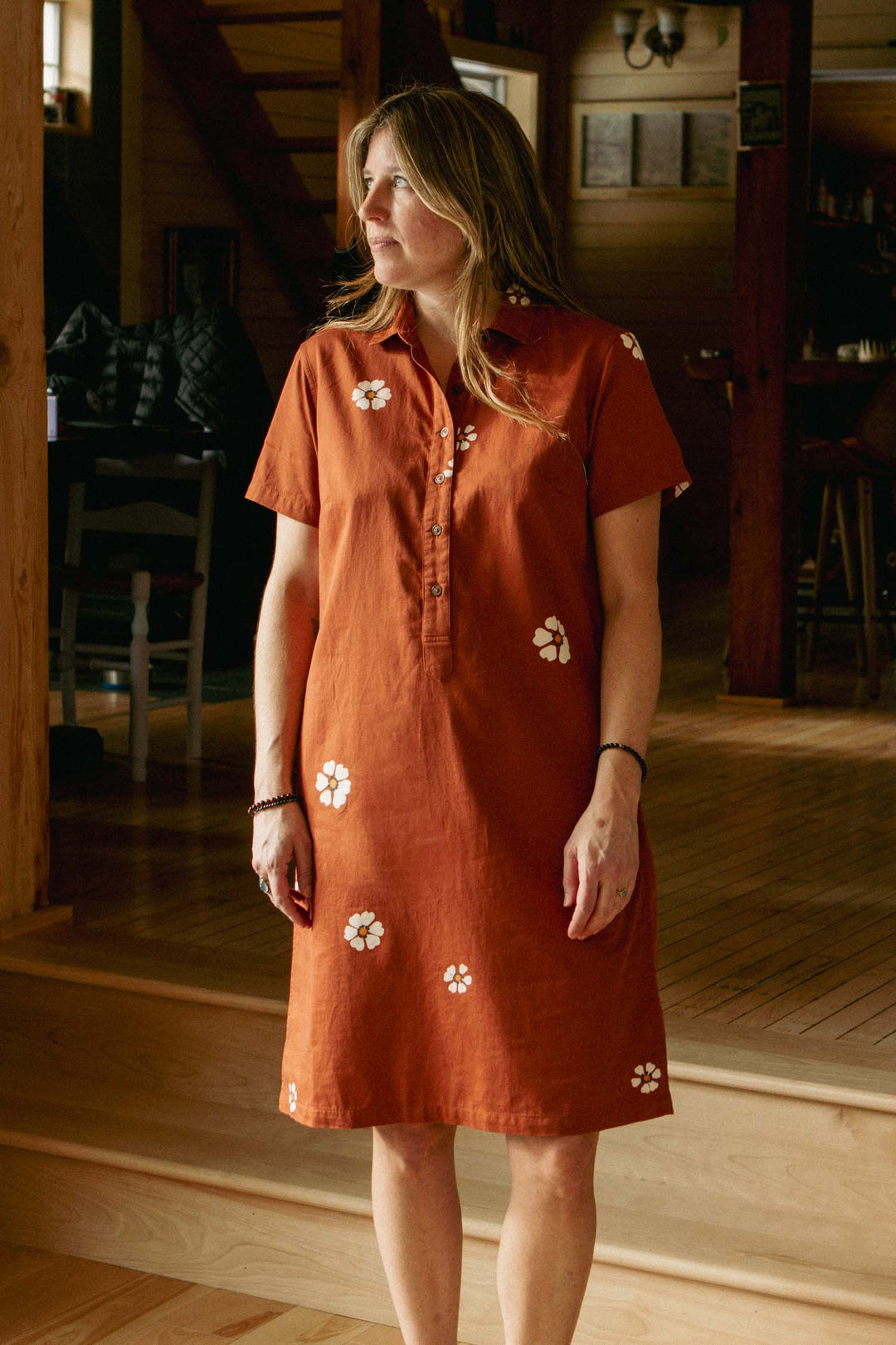 A woman stands indoors on wooden steps, wearing the Bridge & Burn Delia Dress in Redstone Bloom. Warm light highlights her and the floral print dress, with wooden furniture and walls in the background.