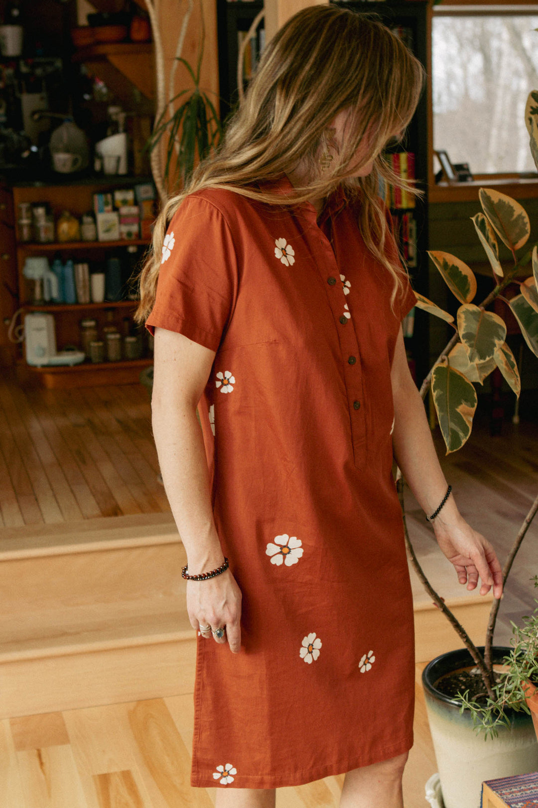 A woman with long hair wears the Bridge & Burn Delia Dress in Redstone Bloom, a rust-colored shirt dress with white floral prints, standing indoors near a potted plant on a wooden floor, looking slightly downward.