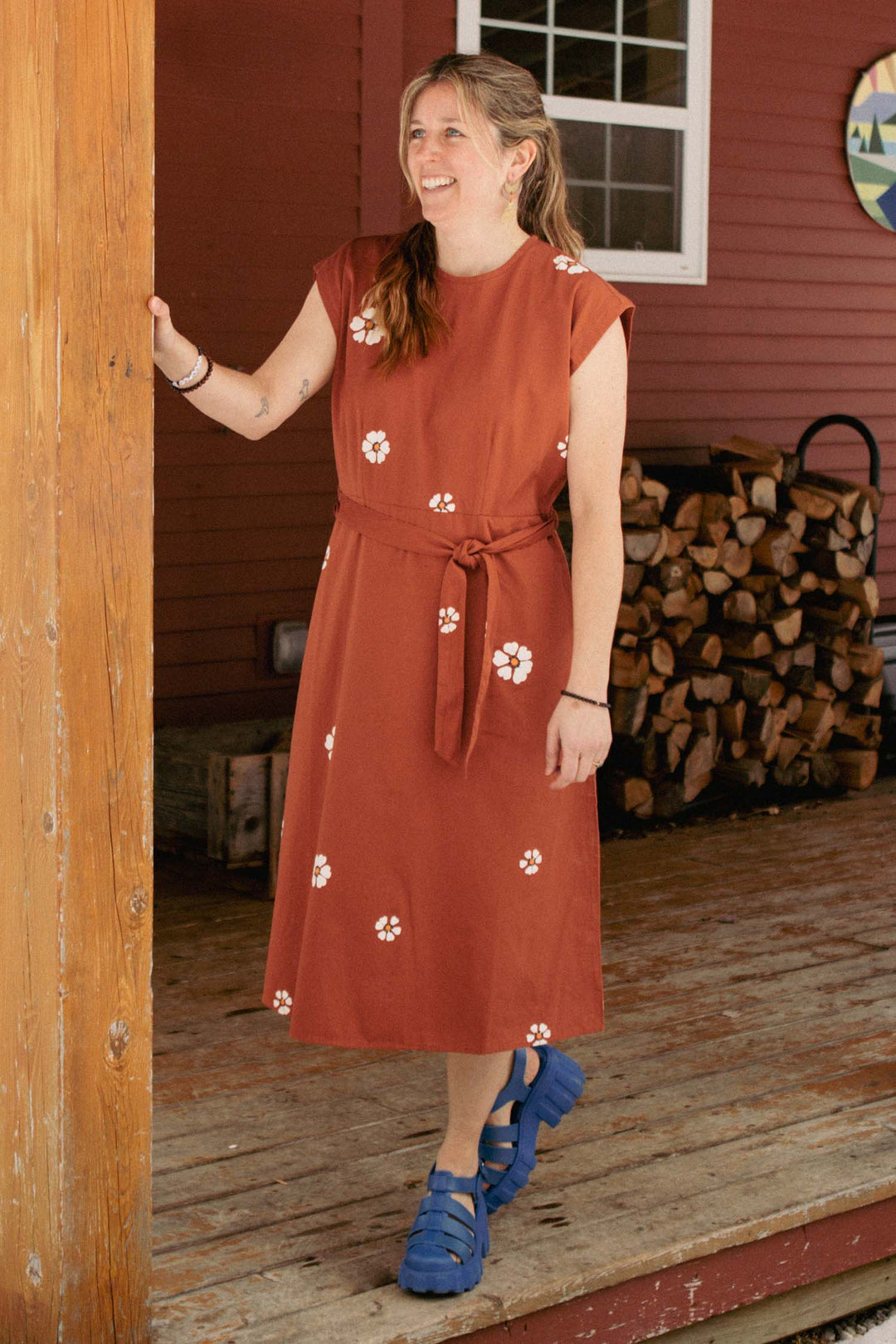 A woman smiles on a wooden porch, wearing the Bridge & Burn Eloise Dress in Redstone Bloom and blue sandals. Stacked firewood and a red building are in the background, highlighting this wardrobe staple.