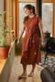 A woman with long hair stands barefoot by a window with potted plants, wearing the Bridge & Burn Eloise Dress in Redstone Bloom. The cotton A-line dress features a tie waist and floral pattern; a wood stove sits in the background.