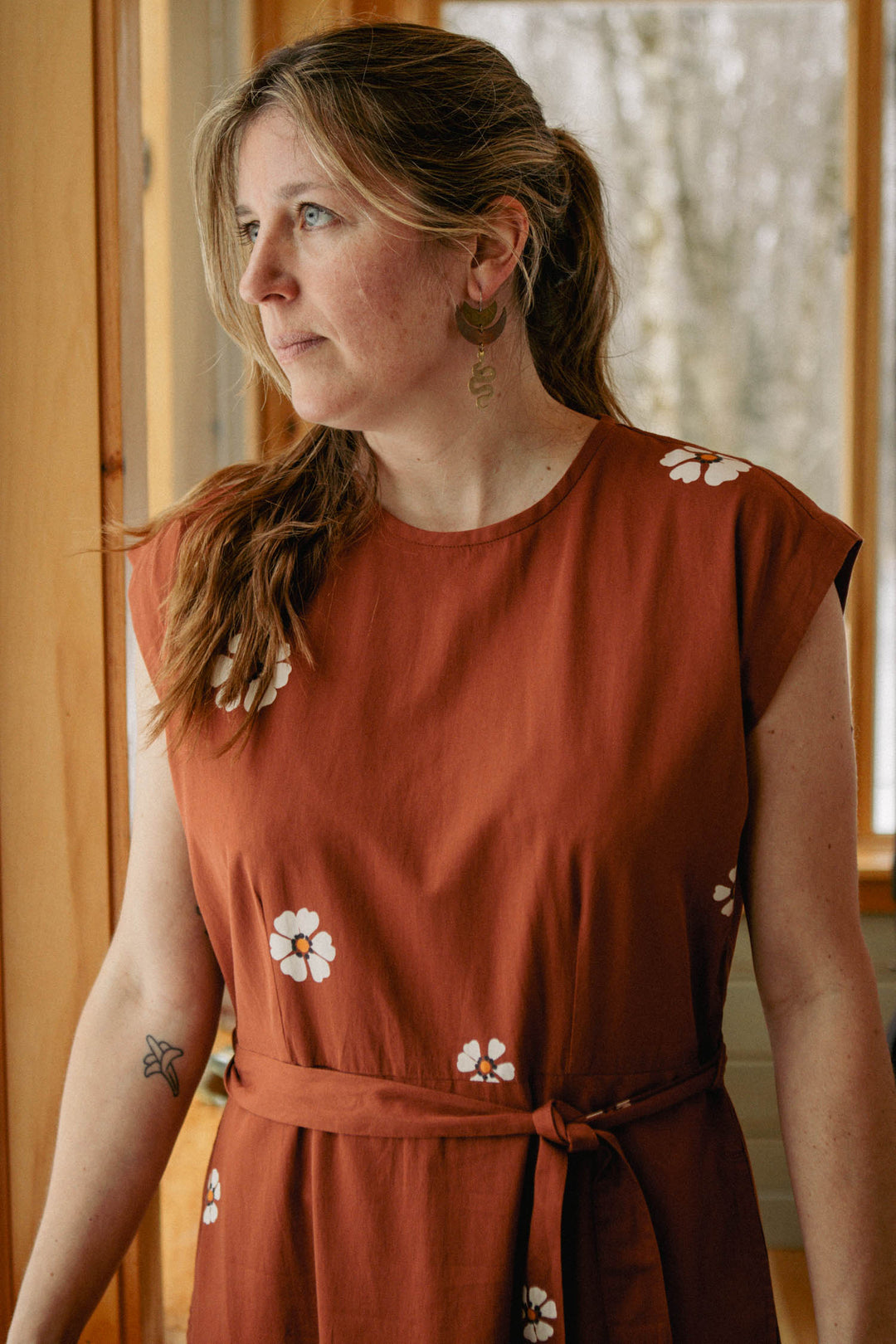 A woman with light brown hair in a ponytail stands by a window, wearing the Bridge & Burn Eloise Dress in Redstone Bloom and gold earrings, looking thoughtfully to the side.