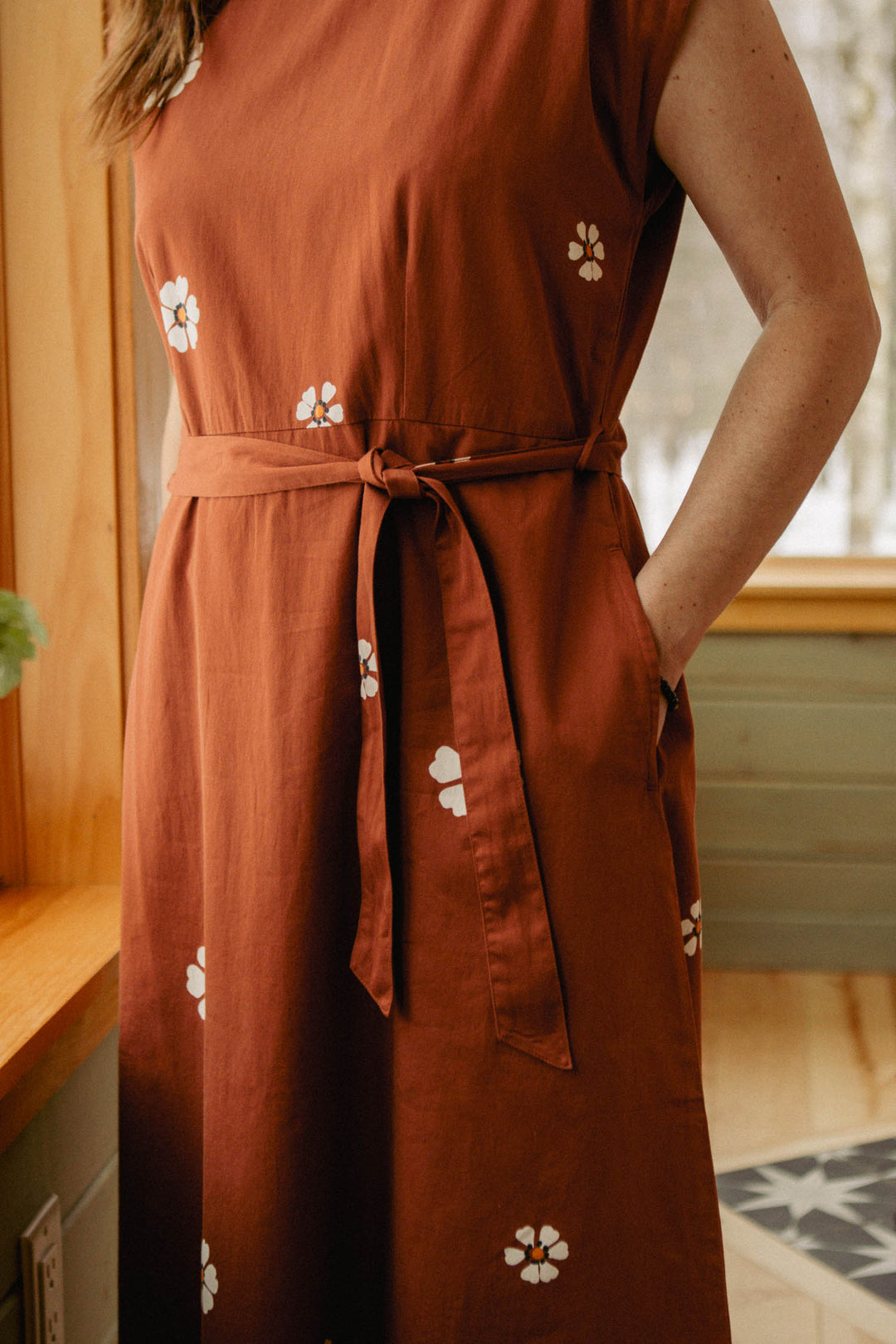 A person stands near a window wearing the Bridge & Burn Eloise Dress in Redstone Bloom—a rust-colored, white floral A-line dress with a waist tie, side pocket, and only the torso and part of one arm visible.