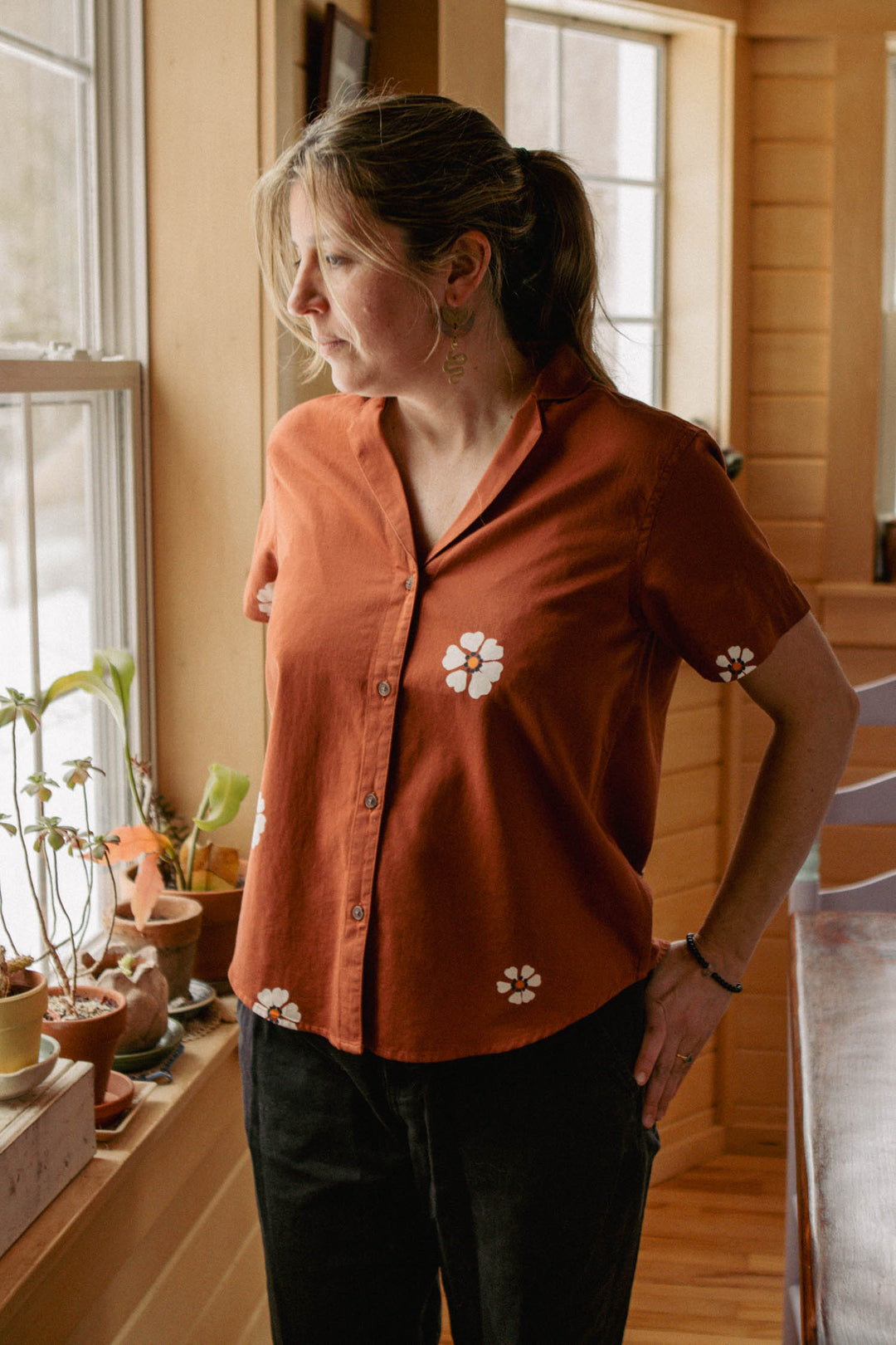 A woman stands indoors by a window with potted plants, wearing the Bridge & Burn Wren Shirt in Redstone Bloom and black pants, enjoying sunlight as it filters into the cozy, wooden room.