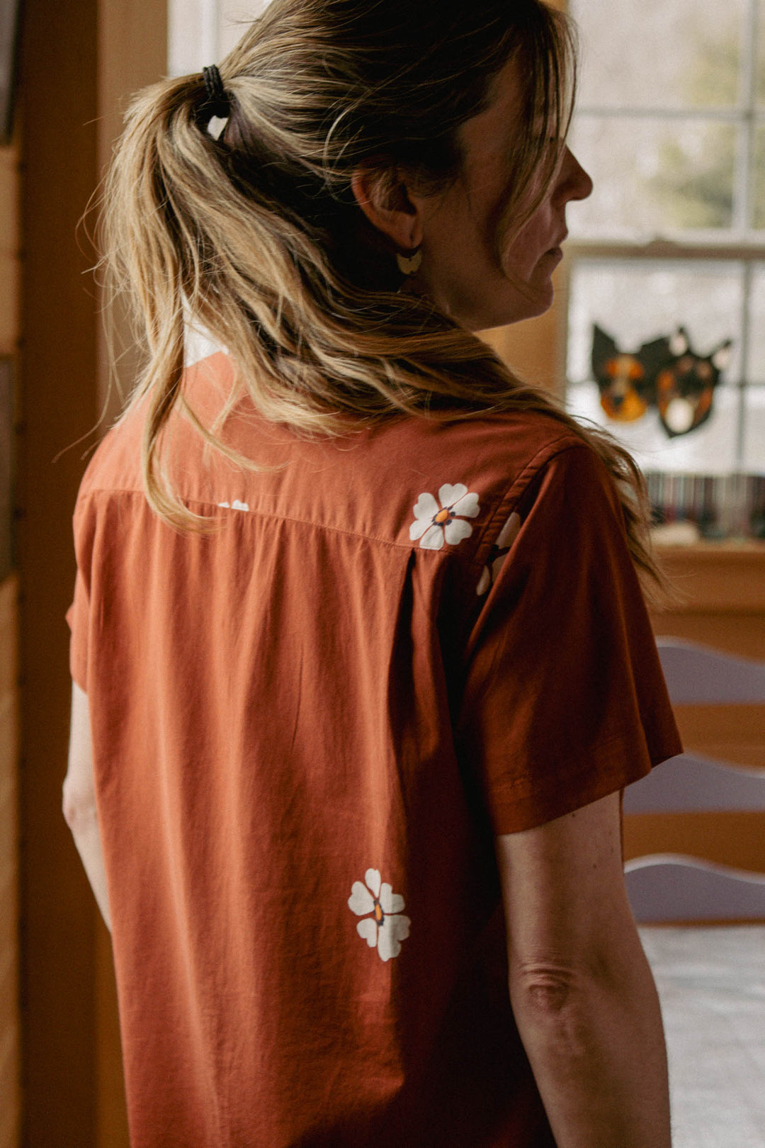 A woman with long, wavy hair in a ponytail stands indoors, sunlight streaming behind her. She wears the Bridge & Burn Wren Shirt in Redstone Bloom—a rust-colored, retro-inspired top with a relaxed fit and white floral pattern.