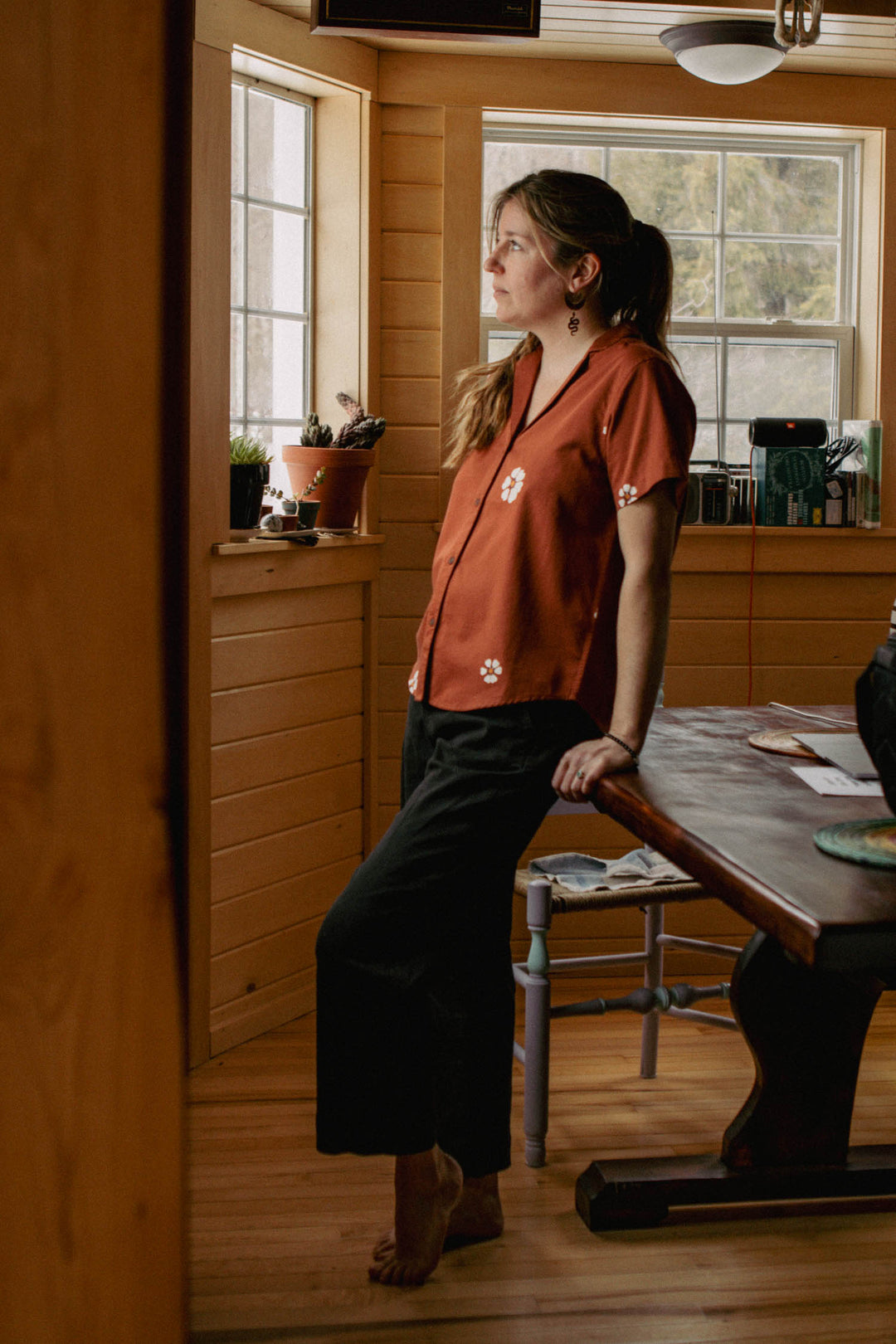 A woman in the Bridge & Burn Wren Shirt in Redstone Bloom and dark pants stands barefoot in a wooden room, leaning against a table as she thoughtfully gazes out the sunlit window, with plants and objects on the windowsill.