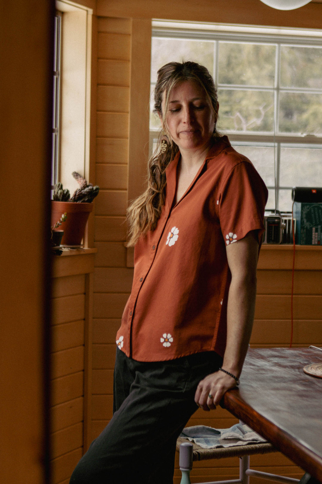 A woman with long hair in a ponytail wears the Bridge & Burn Wren Shirt in Redstone Bloom, a relaxed-fit, retro-inspired top with white floral prints, as she stands by a window in a wooden room, sunlight streaming in.