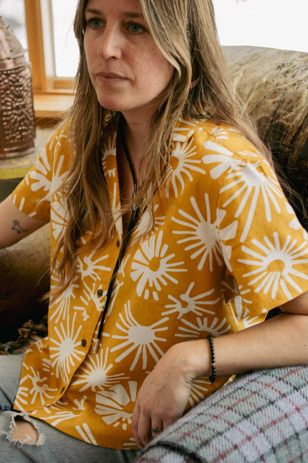 A person with long hair sits on a couch wearing the Bridge & Burn Mila Cropped Shirt in Solar Bloom and ripped jeans, looking slightly to the side with a neutral expression.