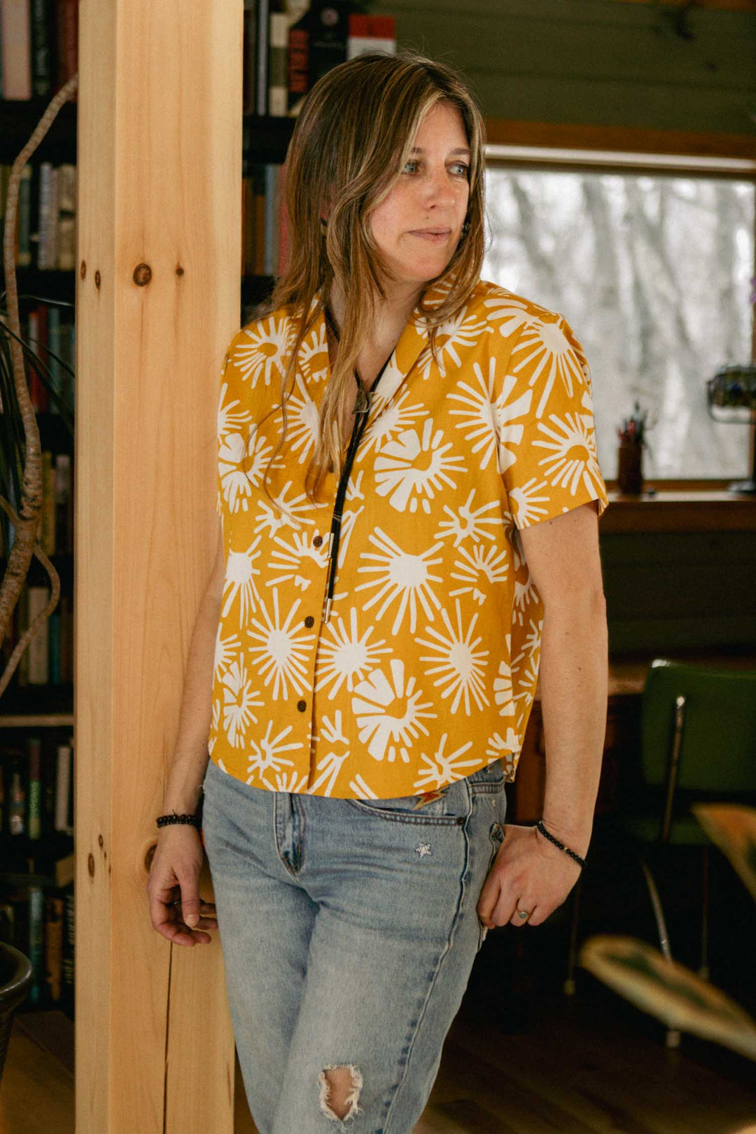 A person with long hair stands indoors by a wooden post, wearing the Bridge & Burn Mila Cropped Shirt in Solar Bloom— a relaxed fit yellow short-sleeve shirt with a white floral pattern. Ripped blue jeans, bookshelves, and a window complete the scene.