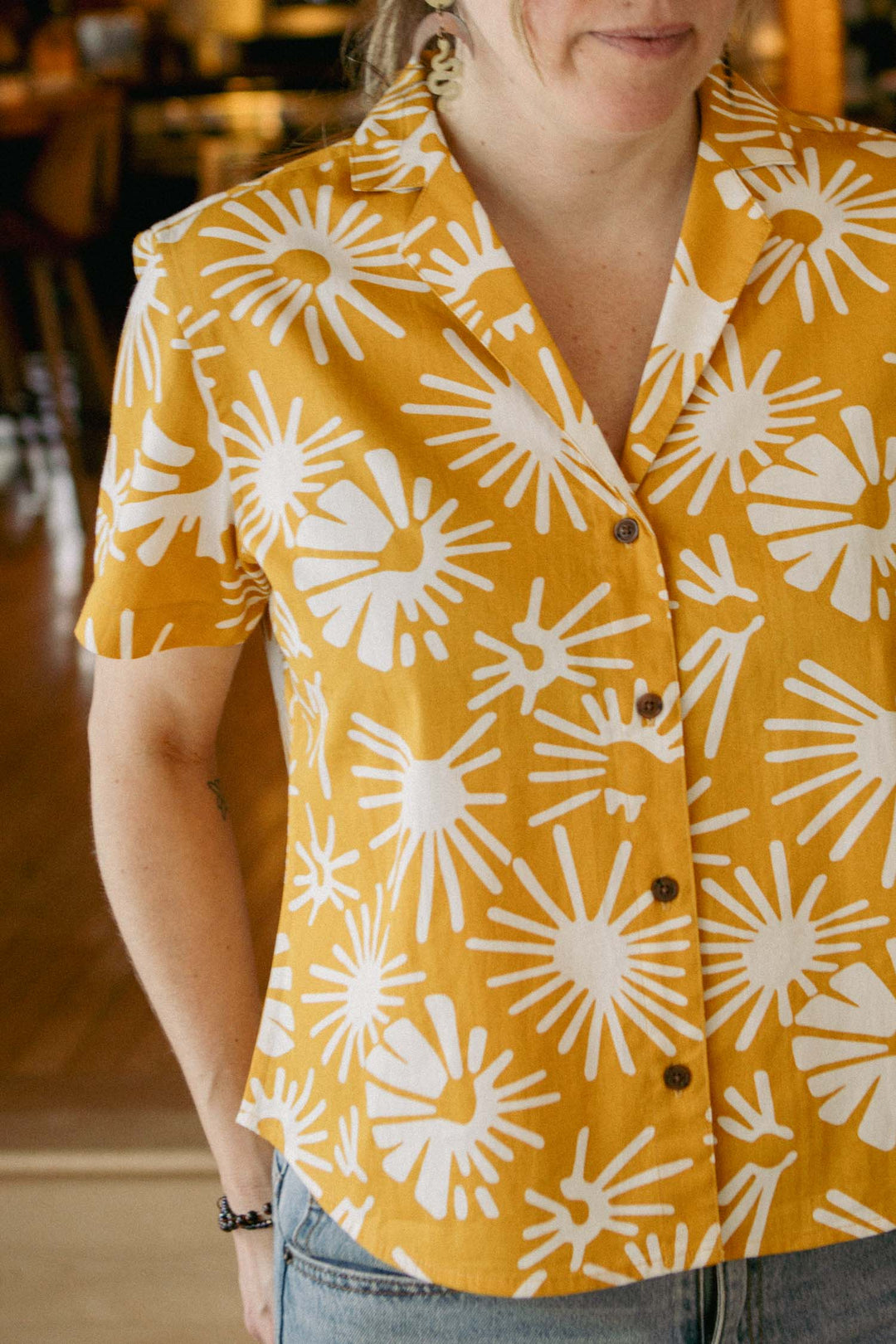 Someone in a Bridge & Burn Mila Cropped Shirt / Solar Bloom—a relaxed fit, short-sleeved, mustard yellow organic cotton shirt with a bold white sunburst print—stands indoors in warm lighting, shown from shoulders to waist.
