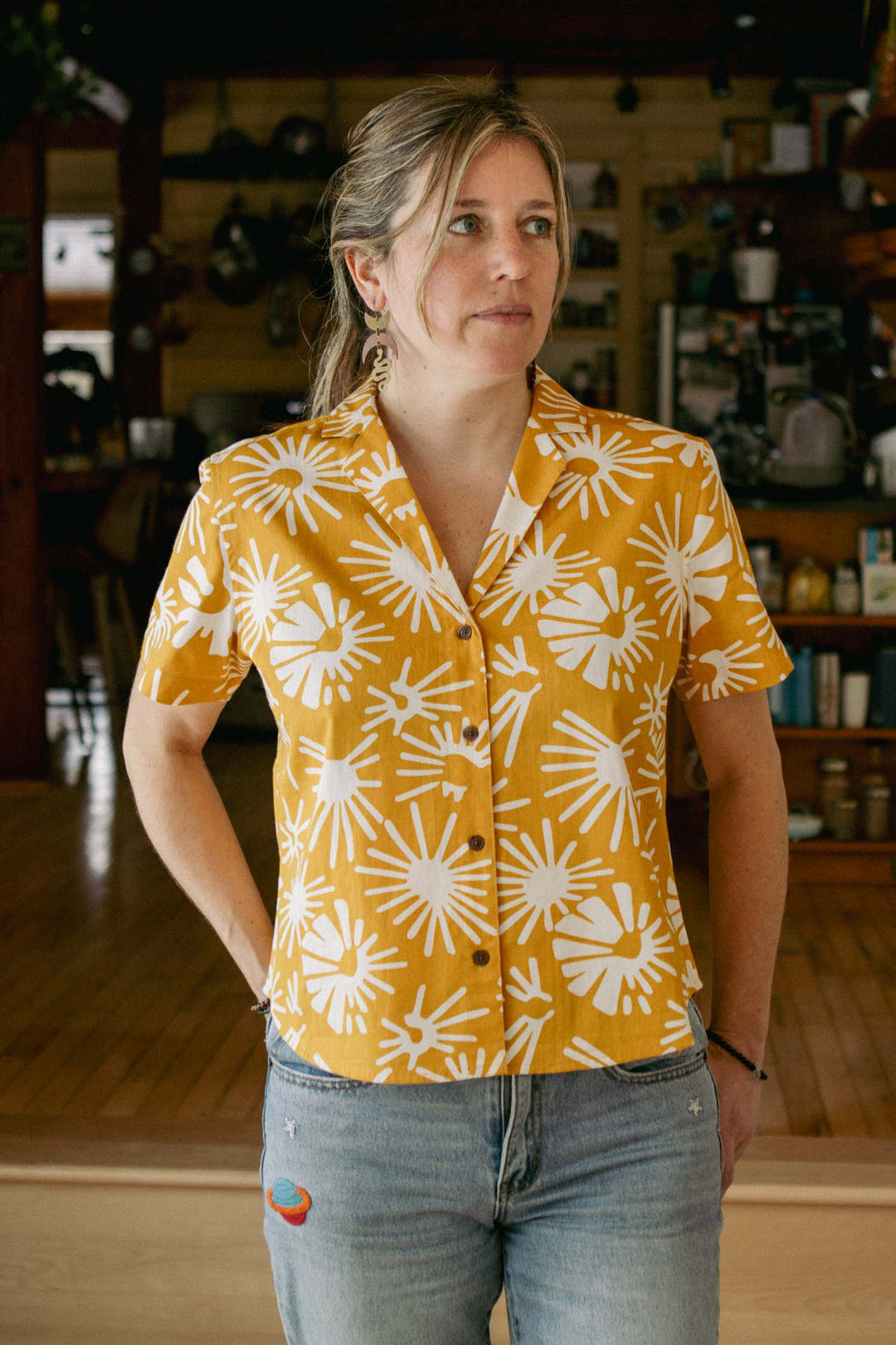 A woman stands indoors wearing the Bridge & Burn Mila Cropped Shirt in Solar Bloom, a relaxed fit yellow short-sleeve top with a bold white floral pattern, paired with light blue jeans. Wooden floors and shelved items are visible in the background.