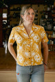 A woman stands indoors wearing the Bridge & Burn Mila Cropped Shirt in Solar Bloom, a relaxed fit yellow short-sleeve top with a bold white floral pattern, paired with light blue jeans. Wooden floors and shelved items are visible in the background.