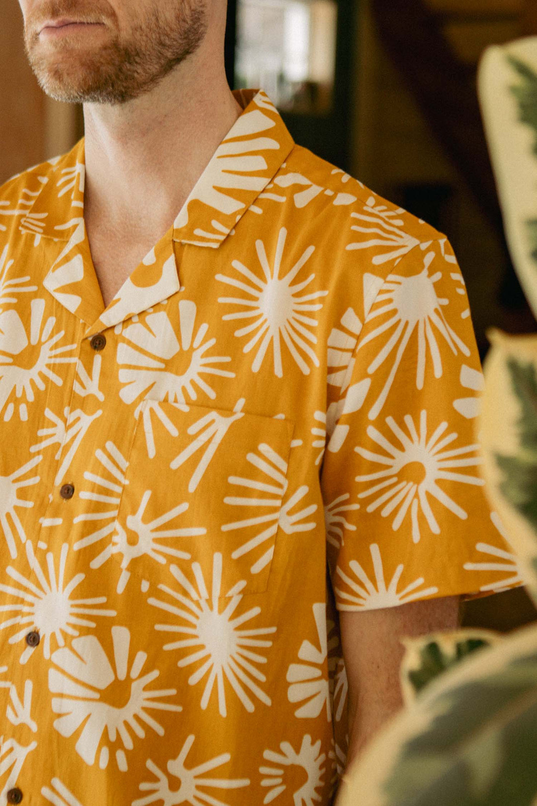 A person wearing the Bridge & Burn Bodie Shirt in Solar Bloom— a retro-inspired, short-sleeved yellow button-up with a bold white sunburst pattern and camp collar—stands indoors near green plants. The image highlights their upper torso and part of their face.