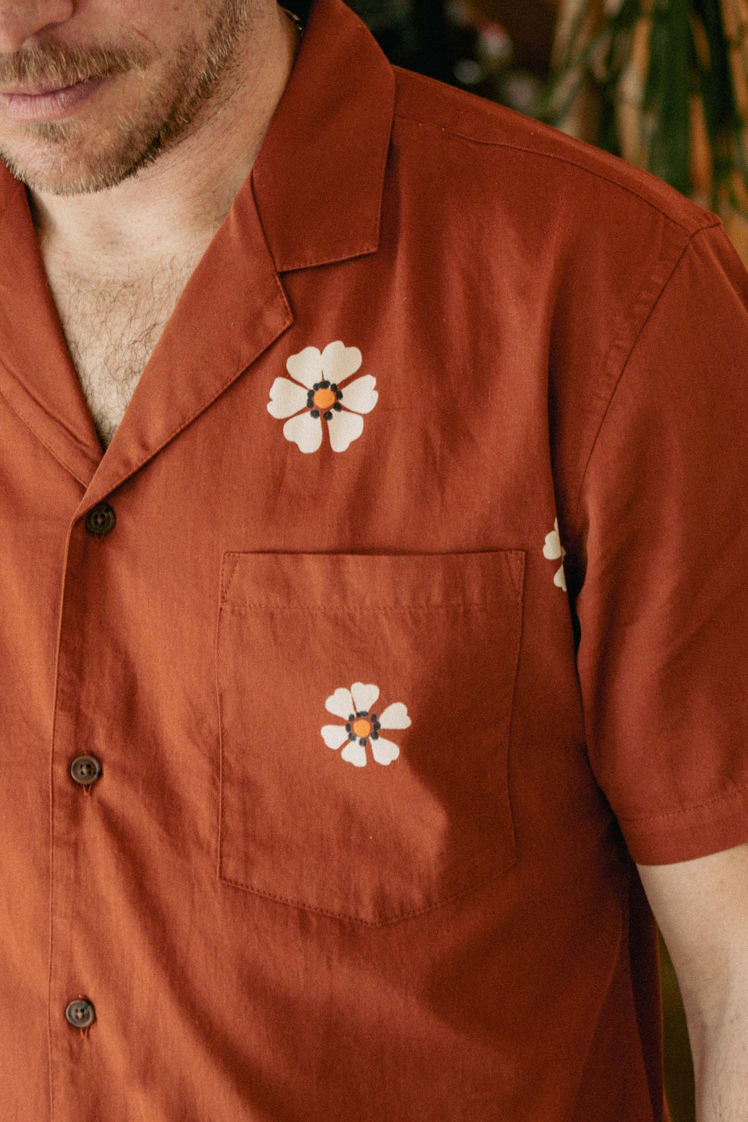 A person wearing the Bridge & Burn Collins Slim Shirt in Redstone Bloom, a rust-colored camp collar button-up with white flower prints above the chest pocket and on the pocket. Only their lower face, chest, and left arm are shown.