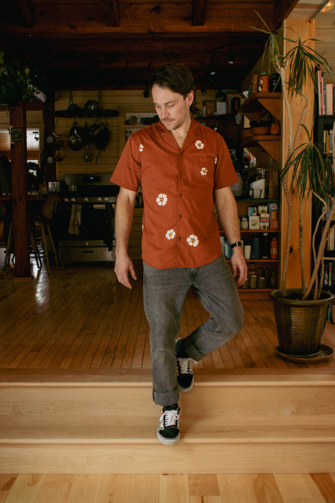 A man wears the Bridge & Burn Collins Slim Shirt in Redstone Bloom and gray slim fit jeans, standing on wooden steps in a cozy, wood-accented kitchen and living space, looking down with one foot on a lower step.