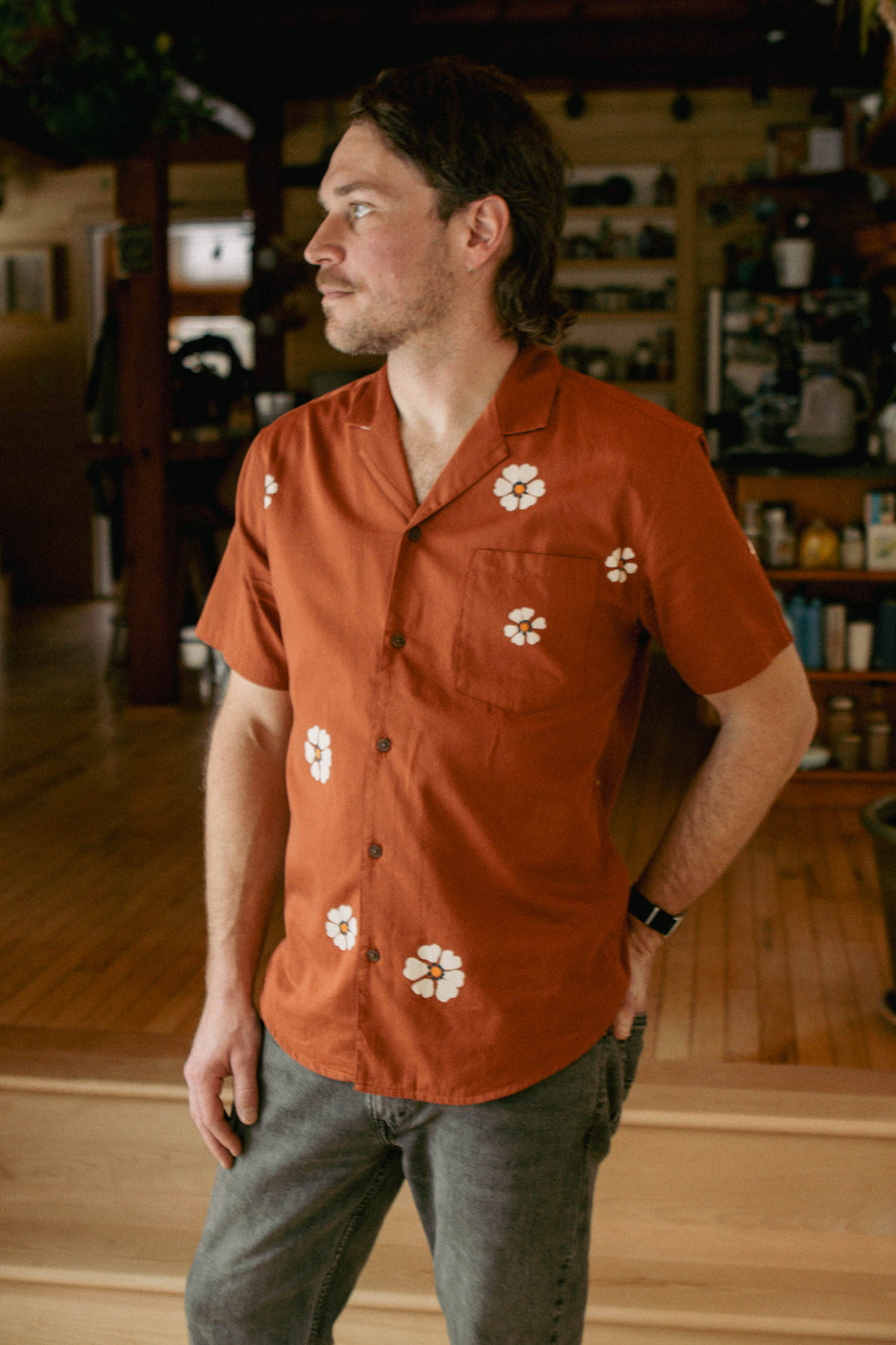 A man with brown hair wears the Bridge & Burn Collins Slim Shirt in Redstone Bloom, featuring a slim fit and white floral print, as he stands indoors on wooden steps looking left among shelves and household items.