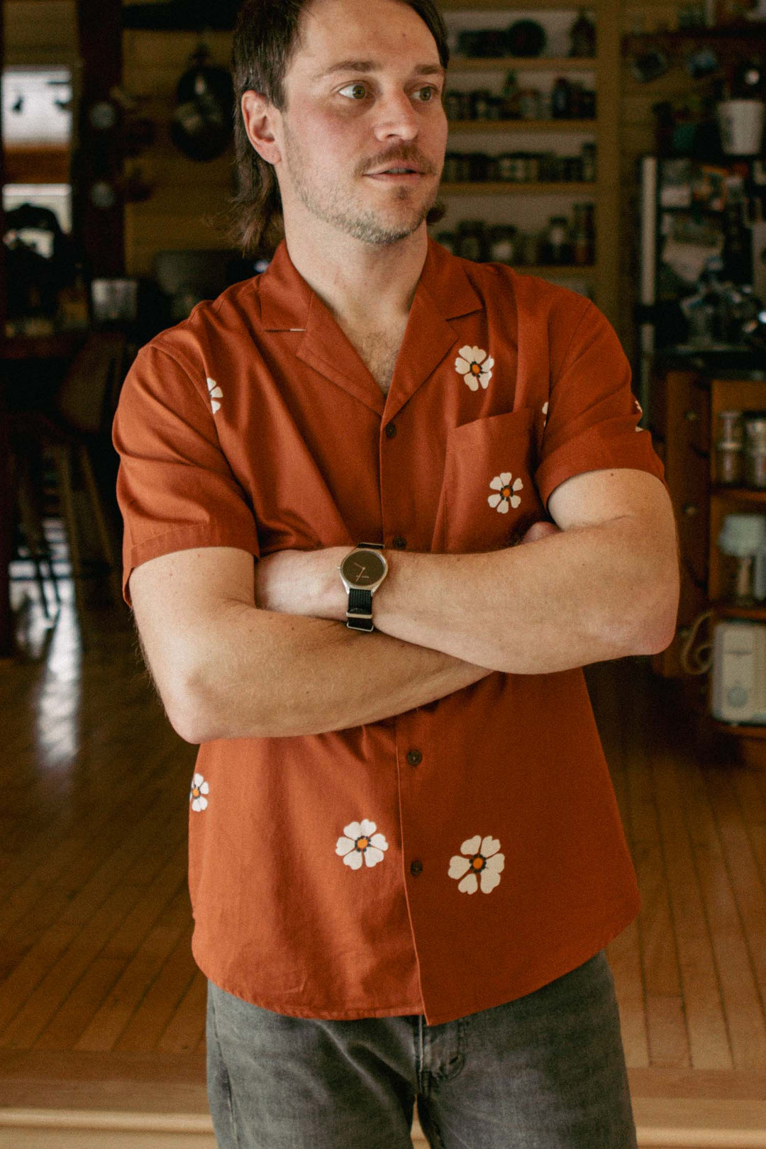 A man with short brown hair and a mustache stands indoors, arms crossed, wearing the Bridge & Burn Collins Slim Shirt in Redstone Bloom—featuring white floral prints—and slim fit gray pants. Wooden floors and shelves fill the background.