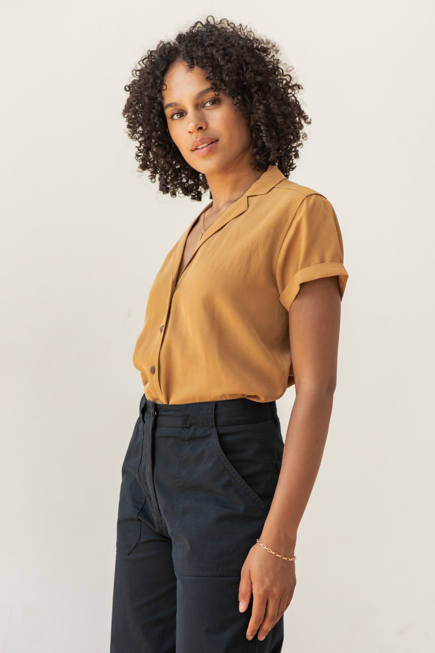 Wearing the Bridge & Burn Innes Shirt in Gold, a person with curly hair and dark high-waisted pants stands against a plain light background, calmly looking at the camera.