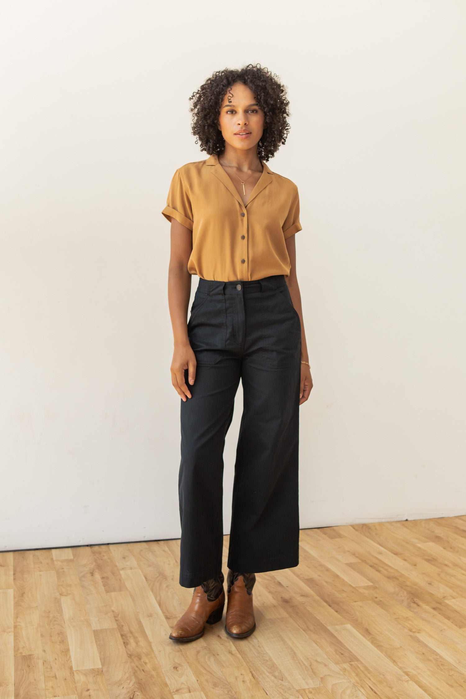 A woman with curly hair stands on a wooden floor against a white wall, wearing the Bridge & Burn Innes Shirt in Gold, high-waisted wide-leg black pants, and brown ankle boots.